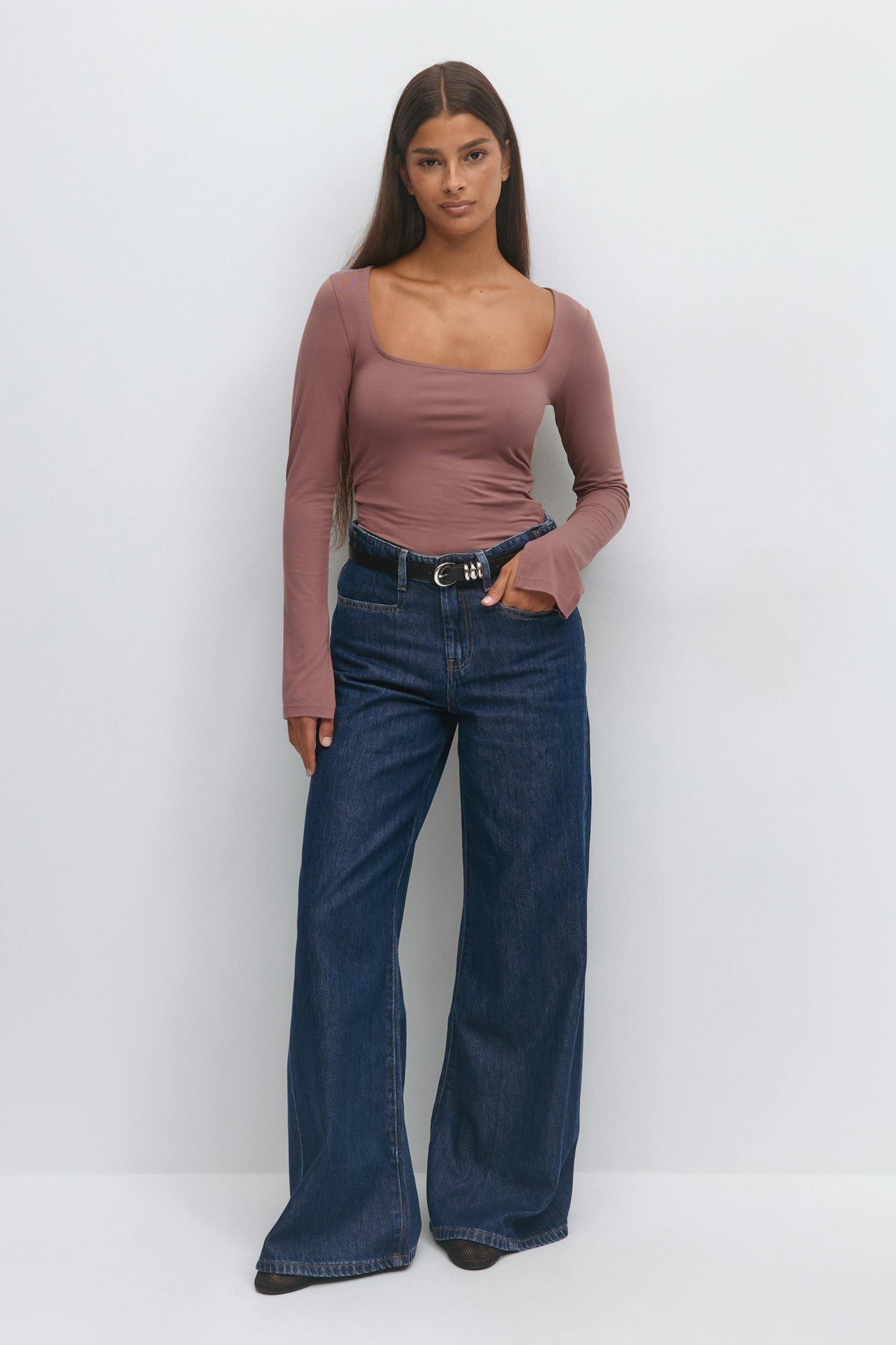 Mauve long-sleeve square-neck fitted top paired with high-waist dark indigo wide-leg jeans, black leather belt with silver double‑ring buckle and pointed black shoes.