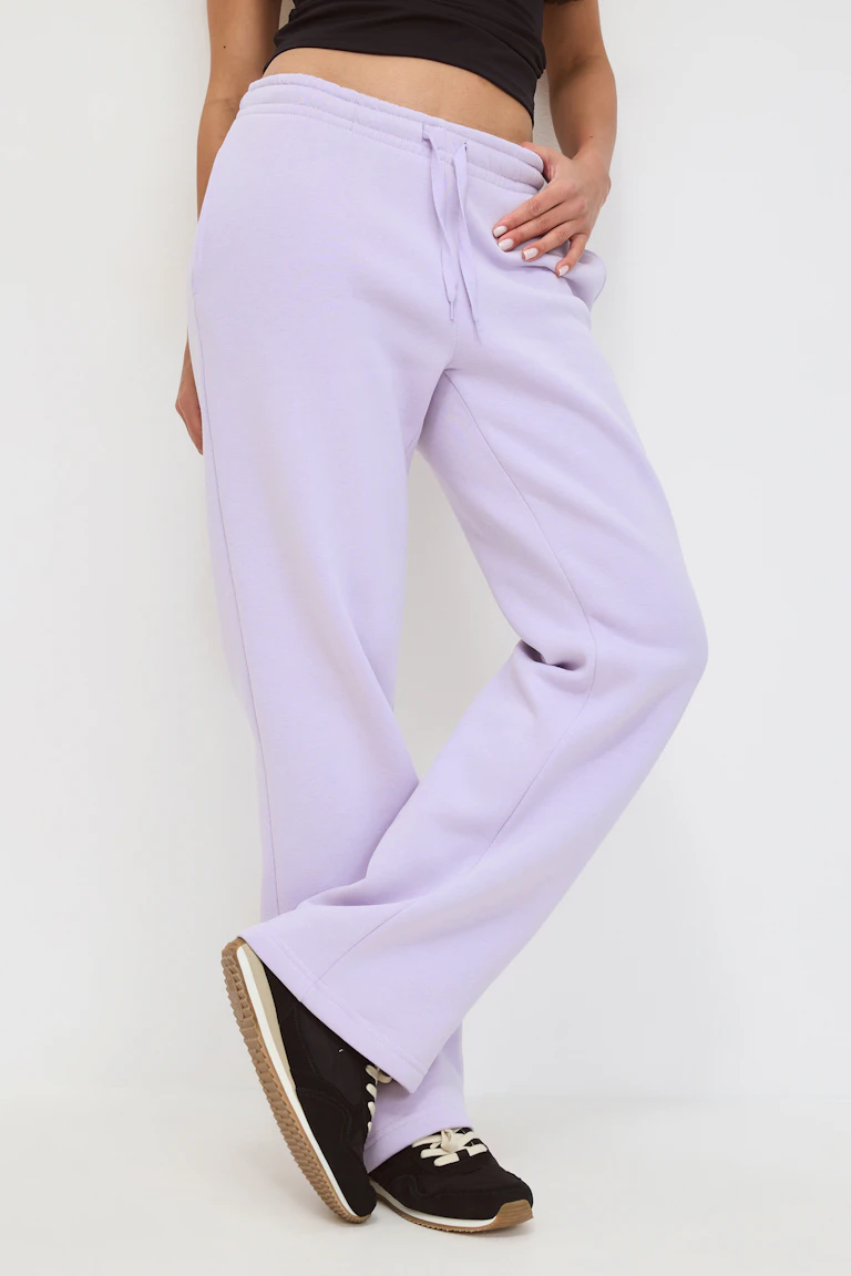 De persoon draagt een licht-lavendelkleurig oversized sweatshirt met capuchon en kangoeroezak en een bijpassende joggingbroek met koord, in laagjes over een wit T-shirt en gecombineerd met bruine veterschoenen.