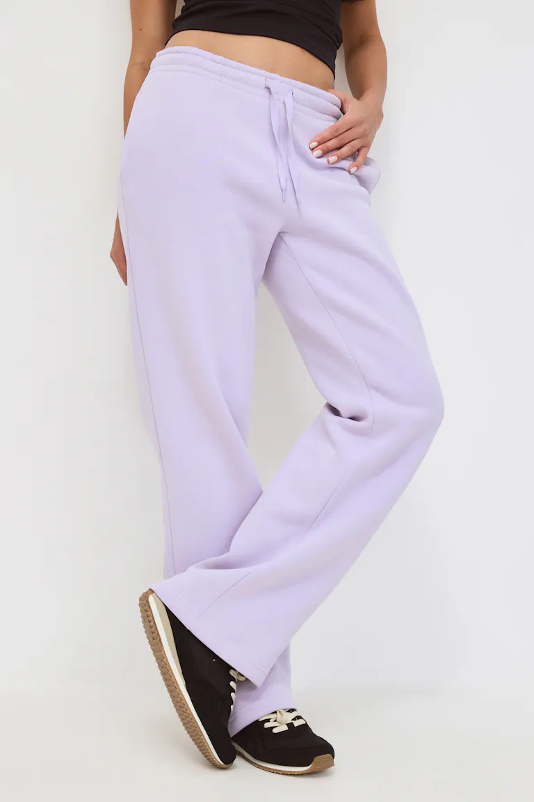 De persoon draagt een licht-lavendelkleurig oversized sweatshirt met capuchon en kangoeroezak en een bijpassende joggingbroek met koord, in laagjes over een wit T-shirt en gecombineerd met bruine veterschoenen.