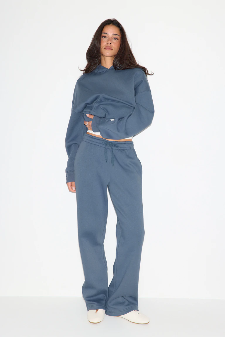 Das Model trägt ein schieferblaues Oversize-Sweatshirt mit Kapuze und eine dazu passende Jogginghose mit Kordelzug, dazu ein weißes, kurz geschnittenes Tanktop, das an der Taille hervorblitzt, sowie weiße Slipper.