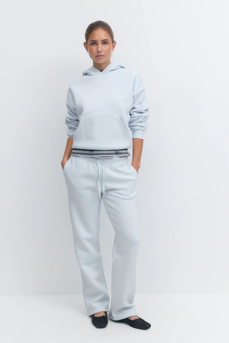 Pale blue relaxed-fit pullover hoodie layered over a striped-hem top, paired with matching pale blue drawstring straight-leg sweatpants and black ballet flats.