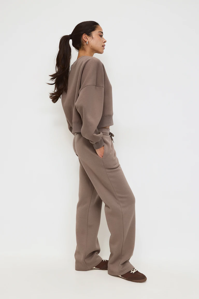 Taupe hooded sweatshirt with kangaroo pocket, matching drawstring relaxed-fit sweatpants, white long-sleeve top with black polka-dot cuffs visible, and black Mary Jane–style ballet flats.