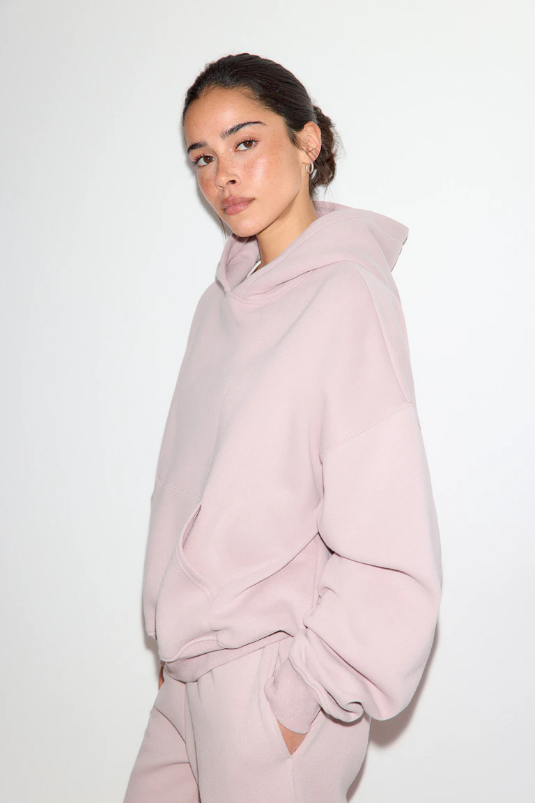 Eine Frau trägt ein hellrosa, übergroßes Kapuzensweatshirt mit Kängurutasche und gerippten Bündchen, dazu eine passende hellrosa Jogginghose.