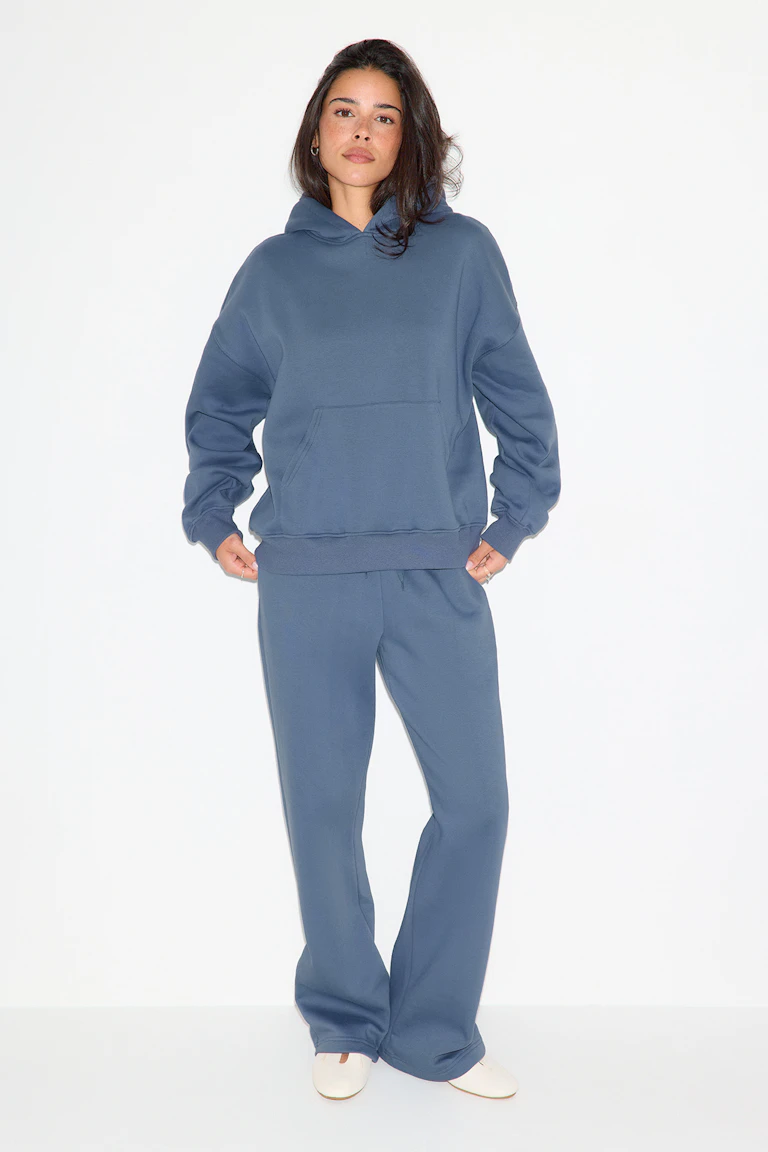 Model wearing a slate-blue oversized hooded sweatshirt with a kangaroo pocket and matching relaxed-fit sweatpants, paired with white slip-on flats.