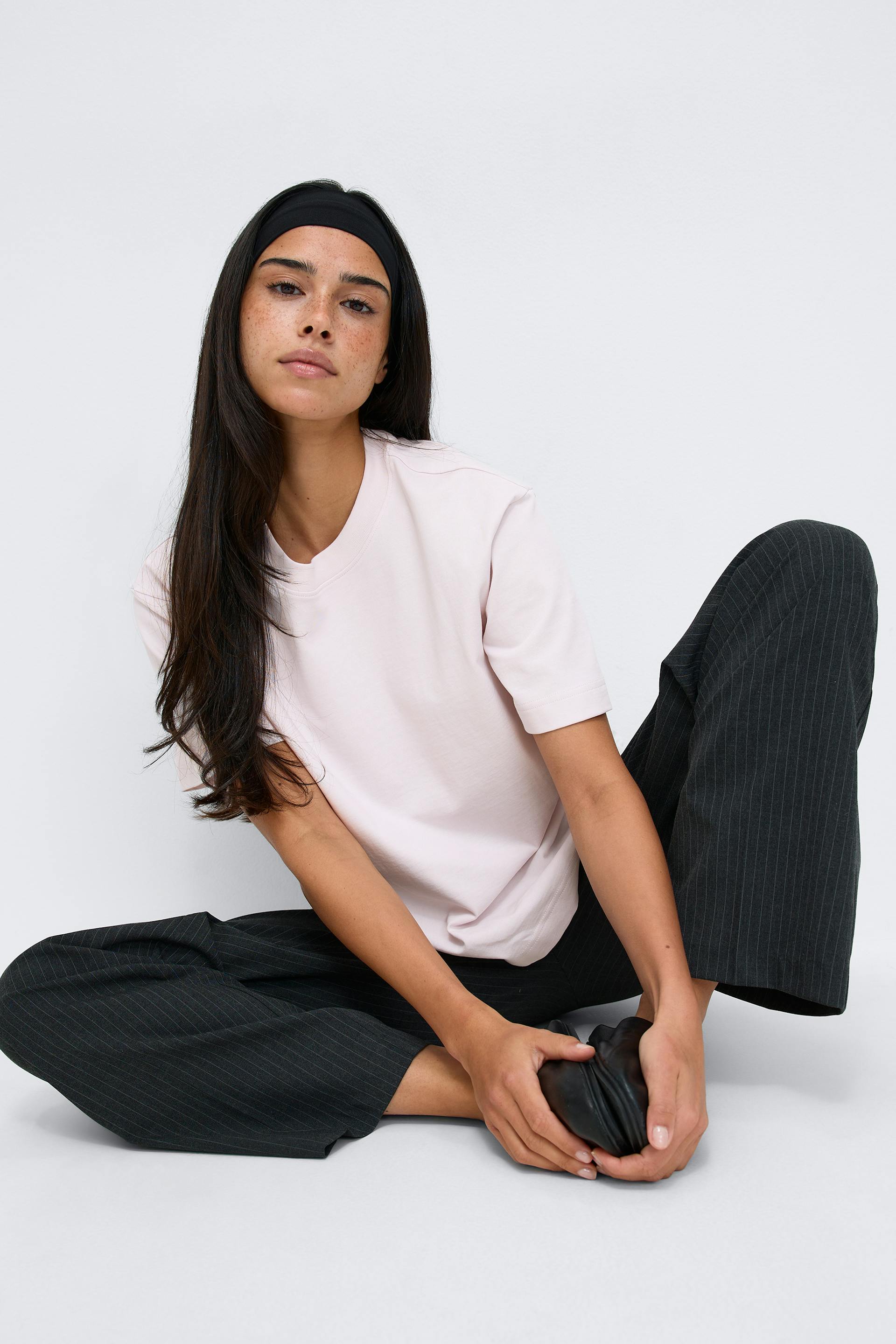 Light-pink crew-neck short-sleeve T‑shirt paired with charcoal pinstripe wide-leg trousers, styled with a black wide headband and a black leather flat held in hand.