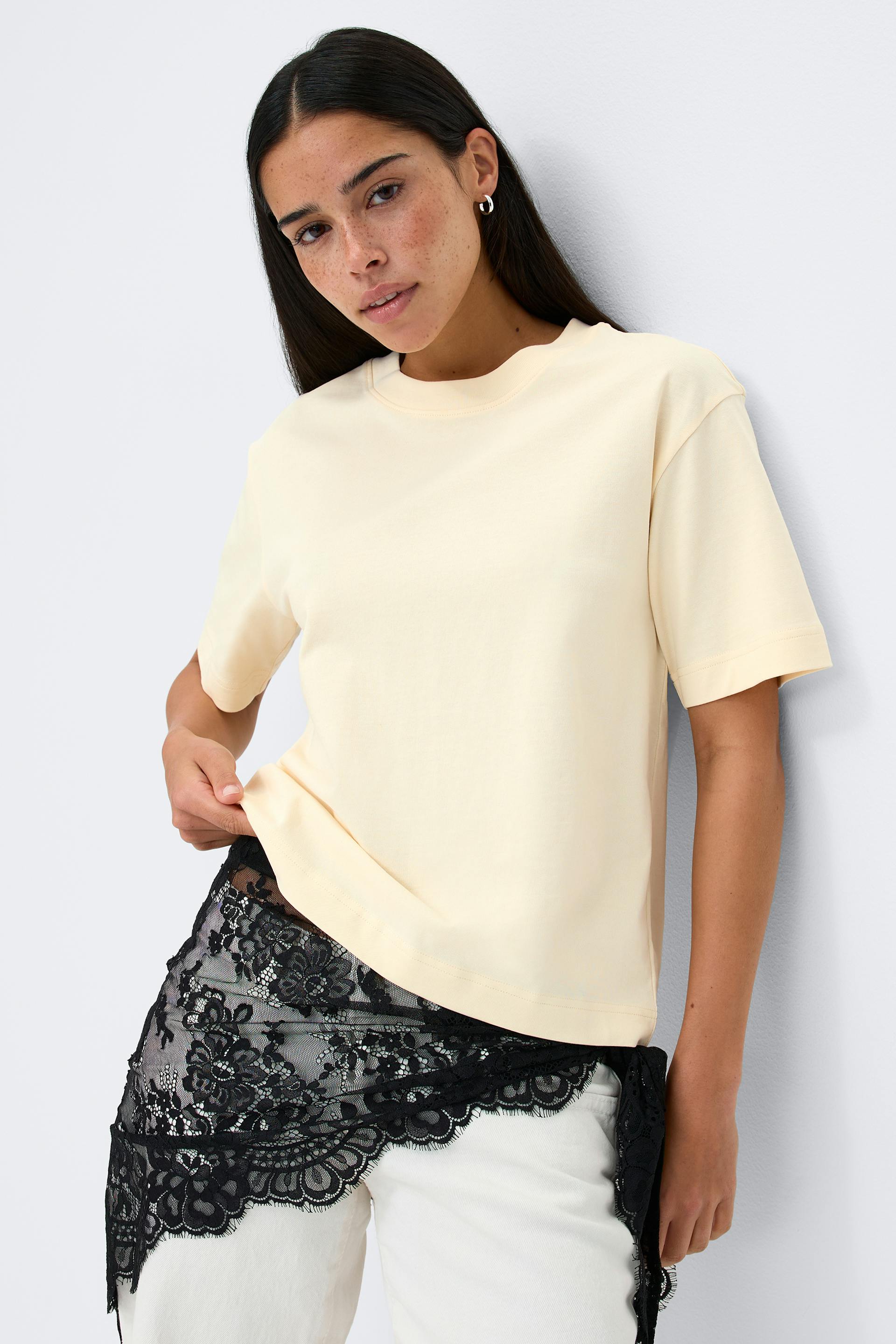Cream relaxed-fit crew-neck short-sleeve T‑shirt paired with white jeans and a black floral lace wrap tied at the hip; small silver hoop earrings.
