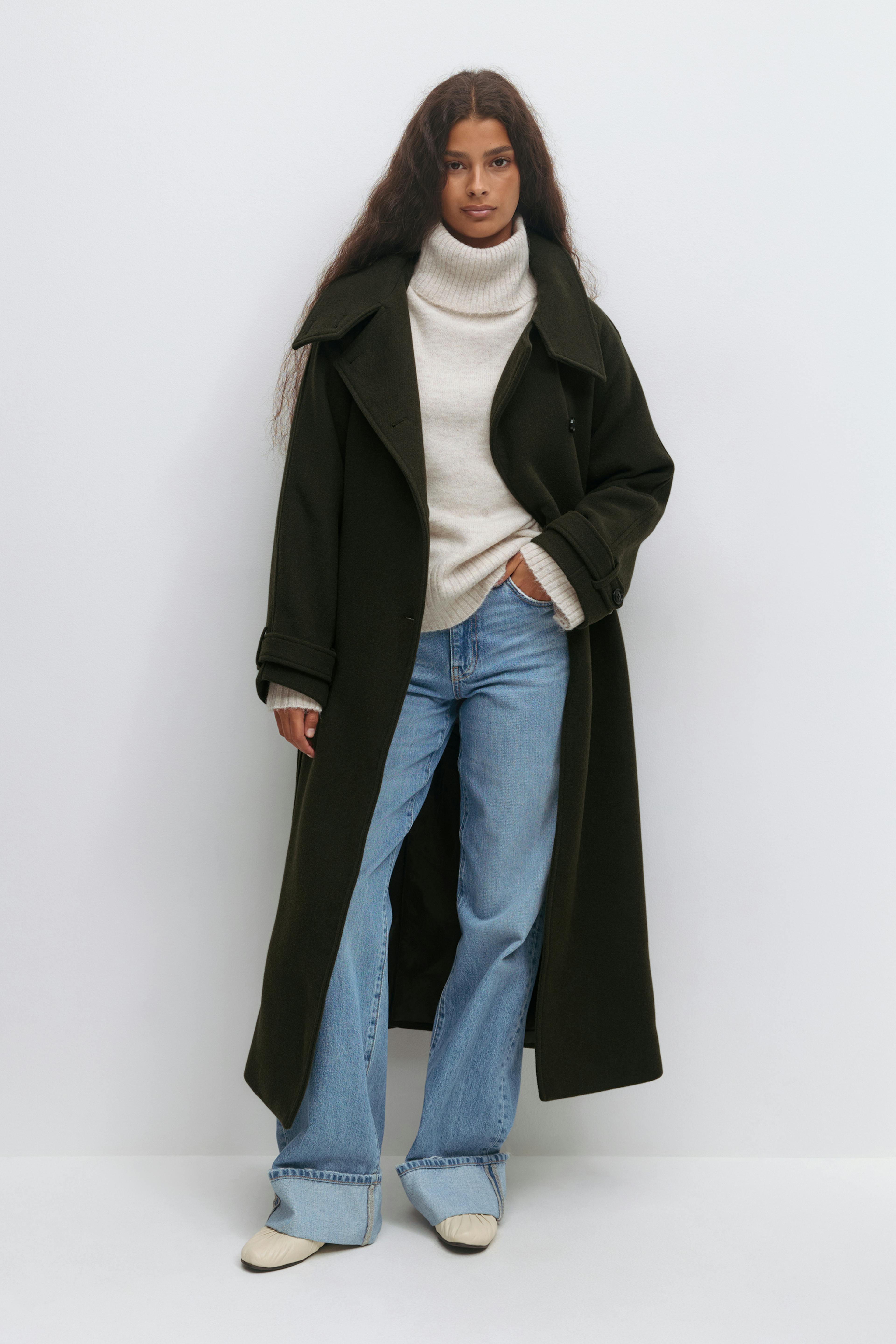 Lange wollen overjas in donkerolijf over een ivoorkleurige geribde coltrui, gecombineerd met een lichtblauwe jeans met wijde pijpen en crèmekleurige platte schoenen.