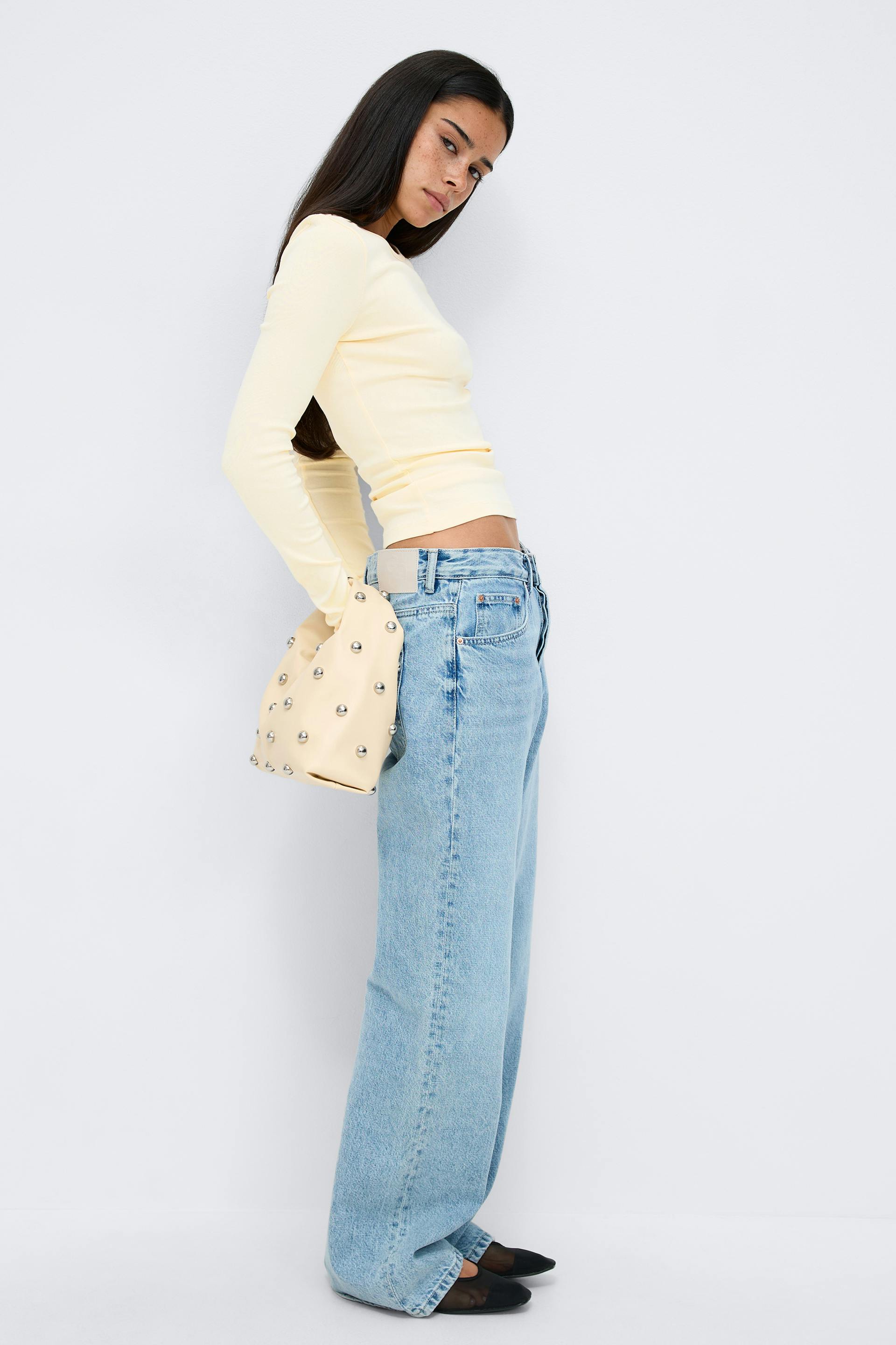 Fitted pale-yellow long-sleeve top, light‑wash high‑rise wide‑leg jeans, cream shoulder bag with silver studs, and black pointed-toe flats.