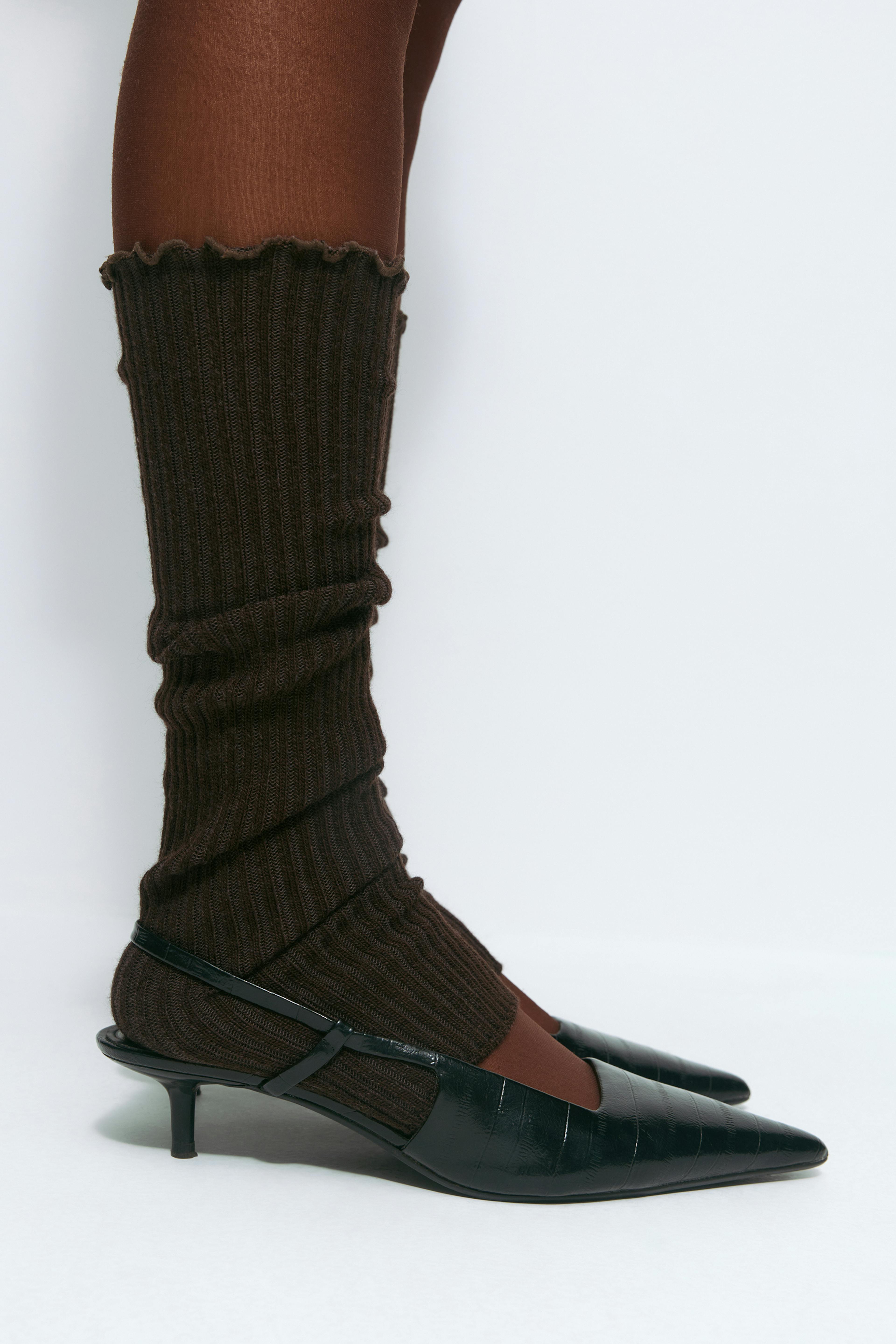 Jambières en maille côtelée marron sur des collants opaques marron, associées à des escarpins à bouts pointus en cuir verni gaufré noir à petits talons.