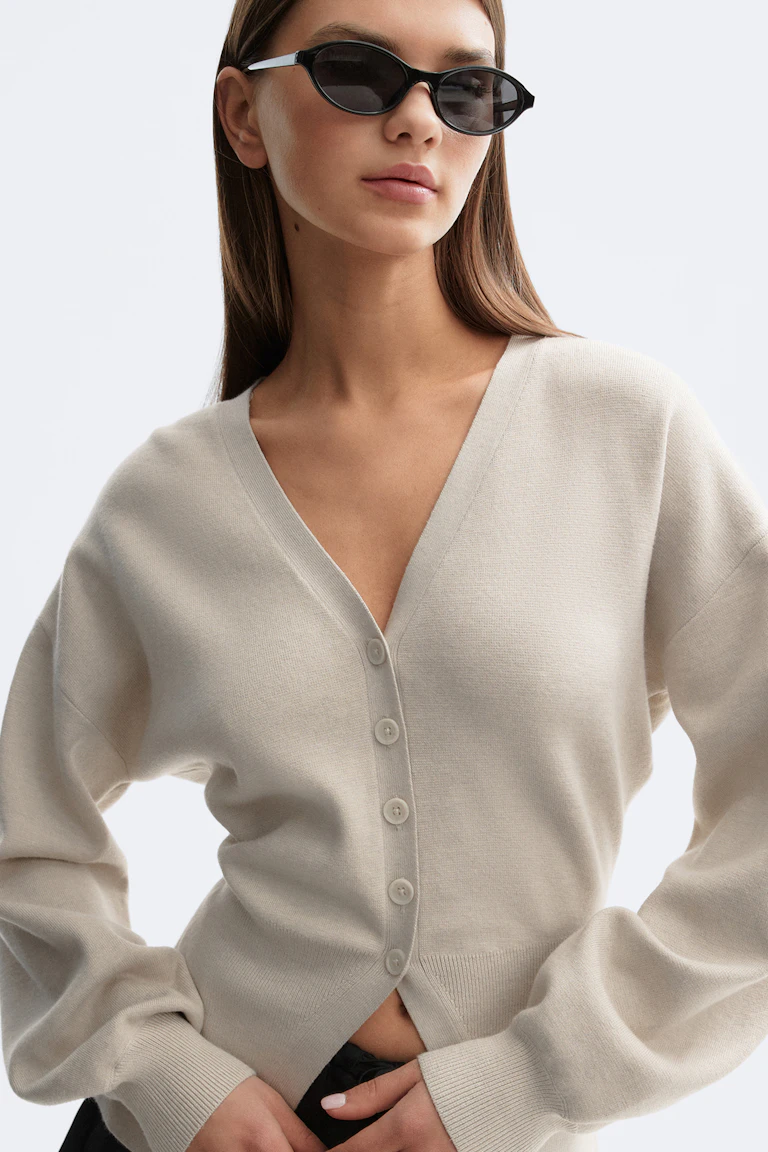 Cream V‑neck button‑front cardigan with long sleeves and ribbed cuffs, paired with slim black sunglasses and black trousers.