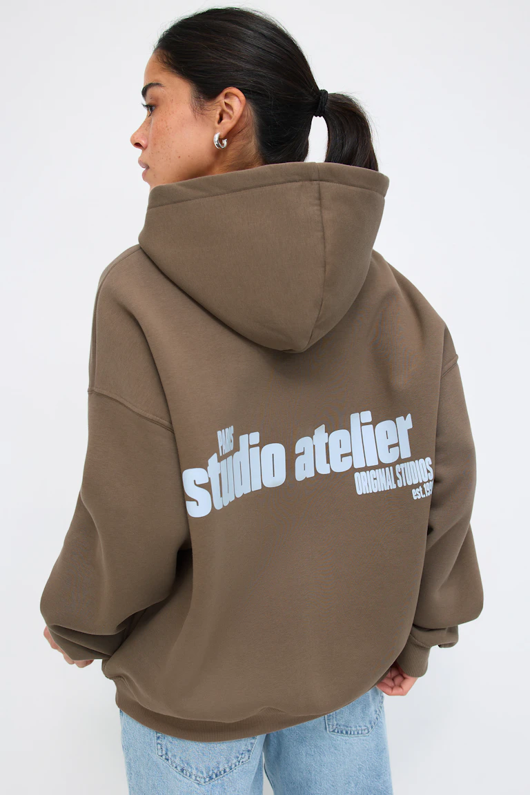 Oversized olijfbruin sweatshirt met capuchon en grote witte "studio atelier"-opdruk op de achterkant, gecombineerd met een jeans van lichtblauw denim.