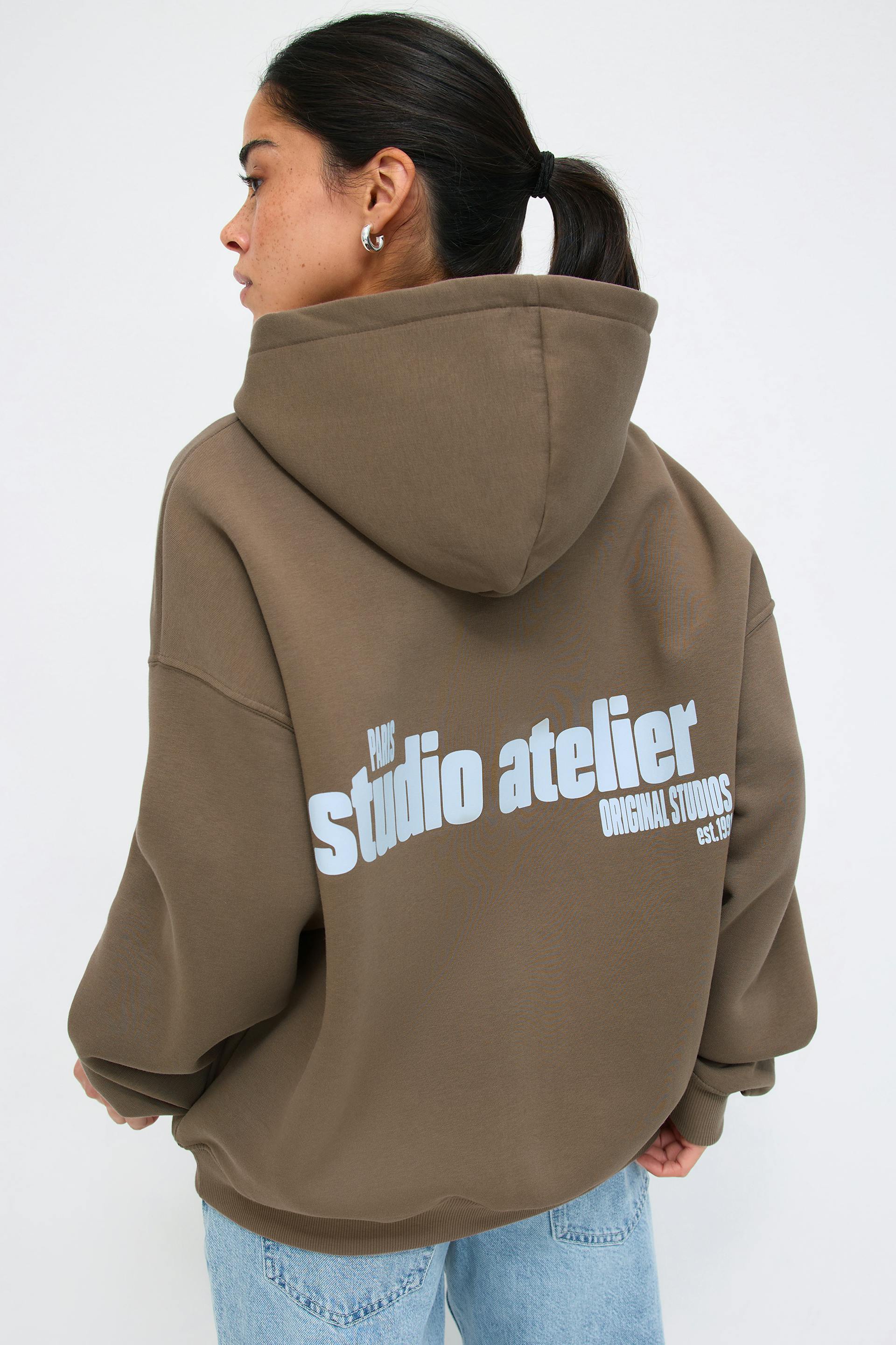 Oversized olive-brown hooded sweatshirt with large white "studio atelier" back print, paired with light-wash blue denim jeans.
