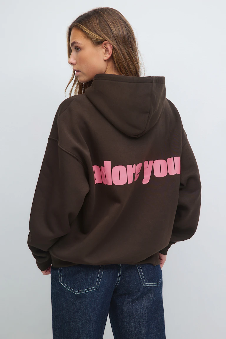 Overdimensjonert, mørkebrun hettegenser med stor, rosa "adore you"-grafikk på ryggen, kombinert med mørkeblå denimjeans.