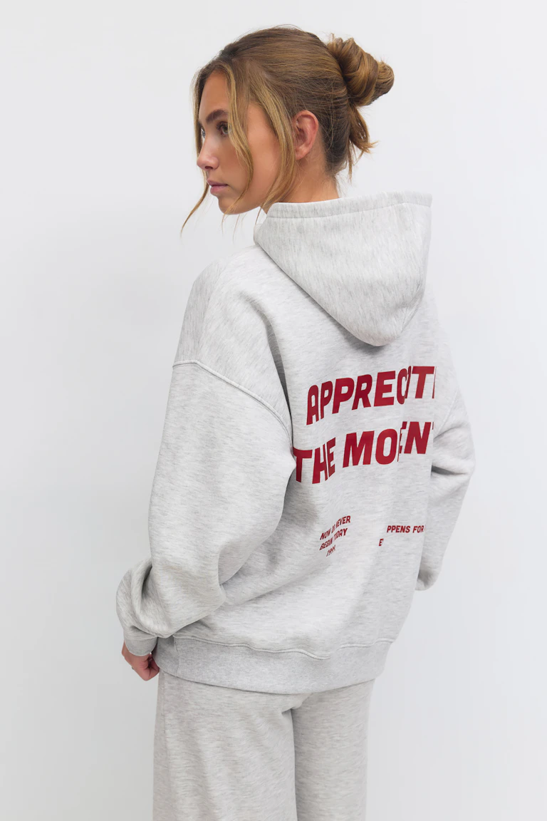 Helles, heidegraues Kapuzensweatshirt in Übergröße mit rotem Blockschriftzug auf dem Rücken, kombiniert mit einer passenden heidegrauen Jogginghose.