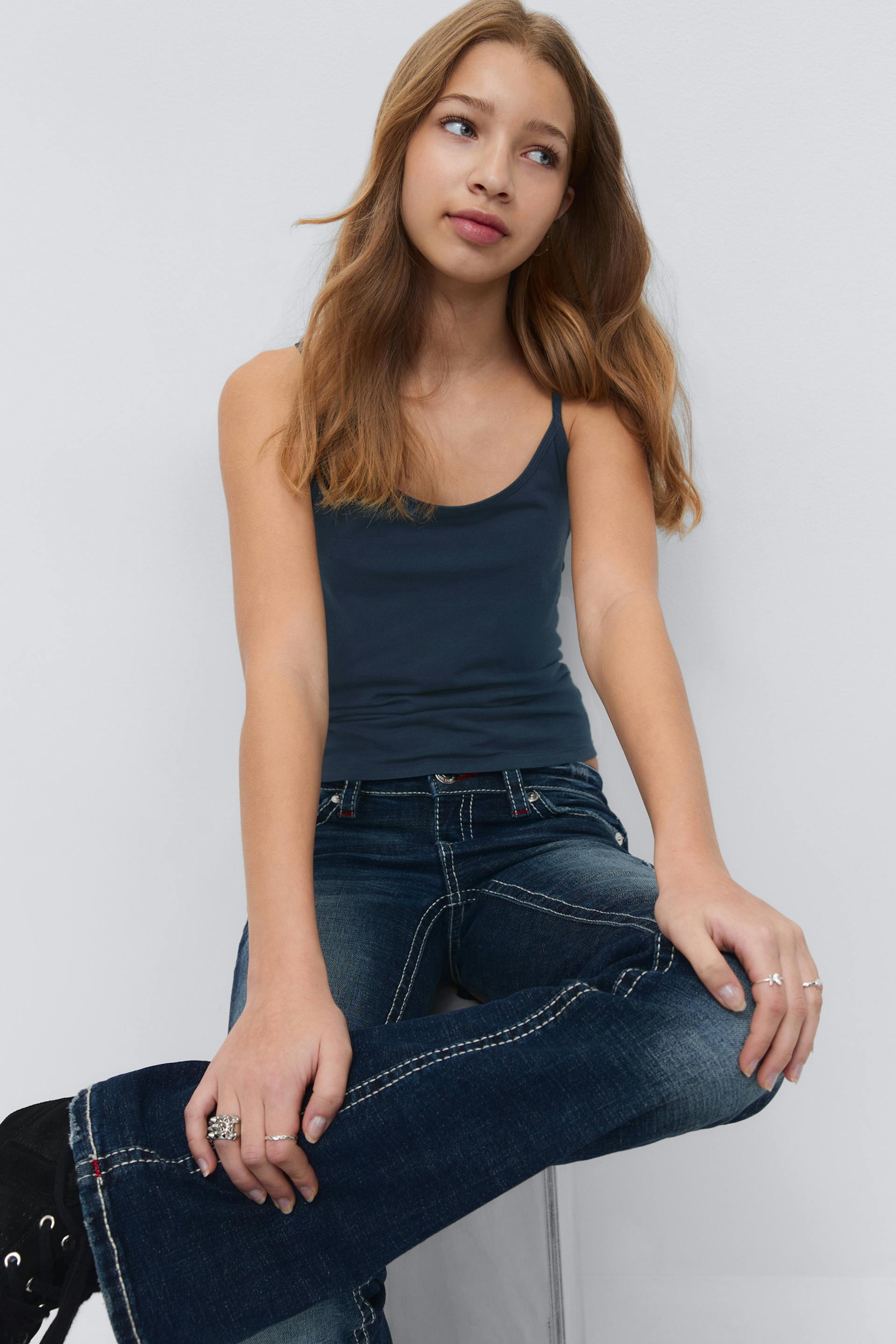 Navy camisole with thin straps, dark-wash jeans with prominent white contrast stitching, black sneakers, and several silver rings.
