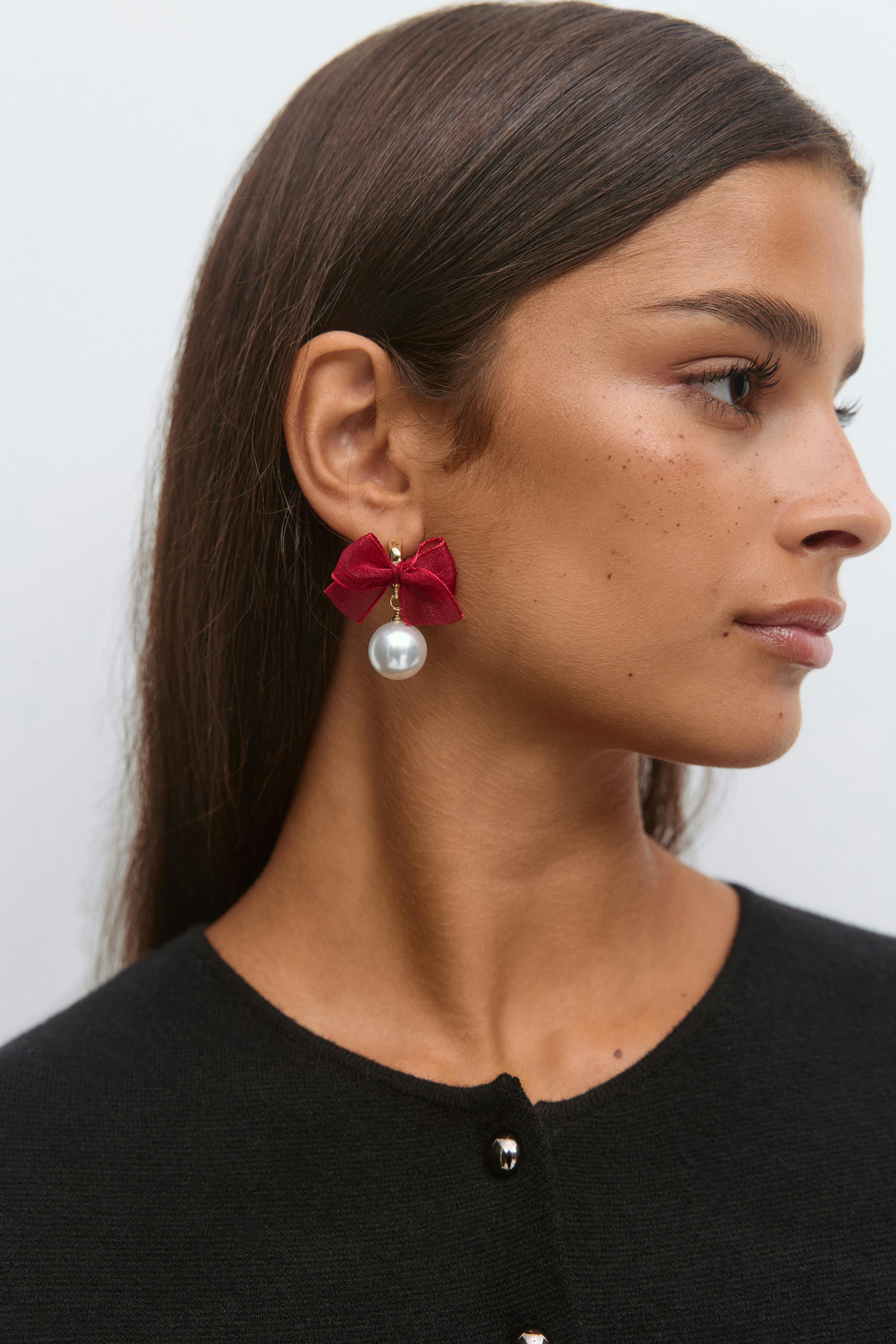 Red satin bow drop earrings with gold fittings and a large white pearl pendant; black round-neck buttoned top.