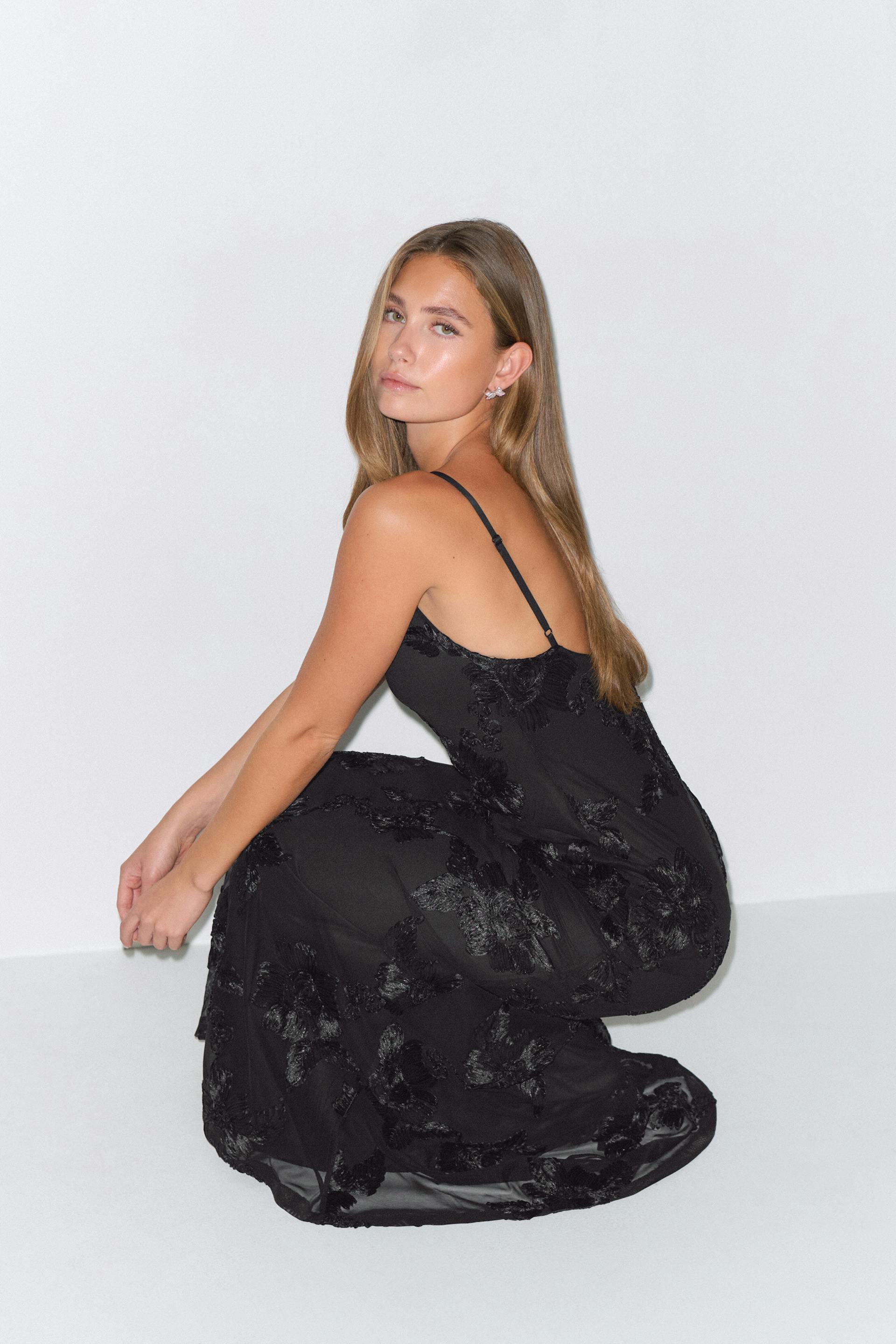 Sort maxikjole med spaghettistropper, strukturerede blomsterapplikationer og halvgennemsigtig kant.