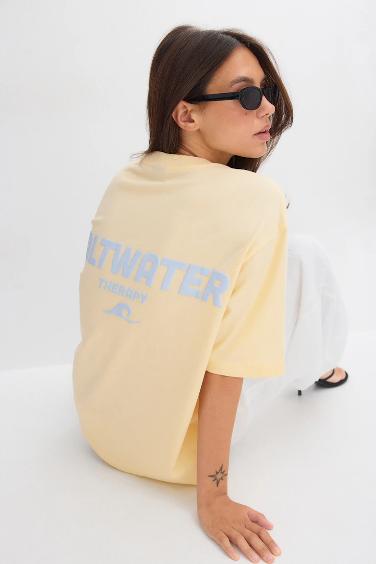T-shirt surdimensionné jaune pâle avec imprimé "SALTWATER THERAPY" bleu clair au dos ; pantalon large blanc ; lunettes de soleil ovales étroites noires ; sandales à talons noires.