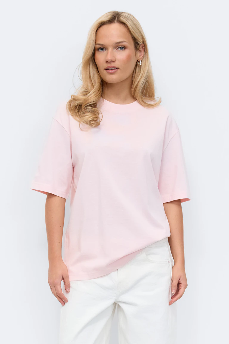 Light pink oversized short-sleeve crewneck T-shirt paired with white relaxed-fit trousers and small gold hoop earrings.