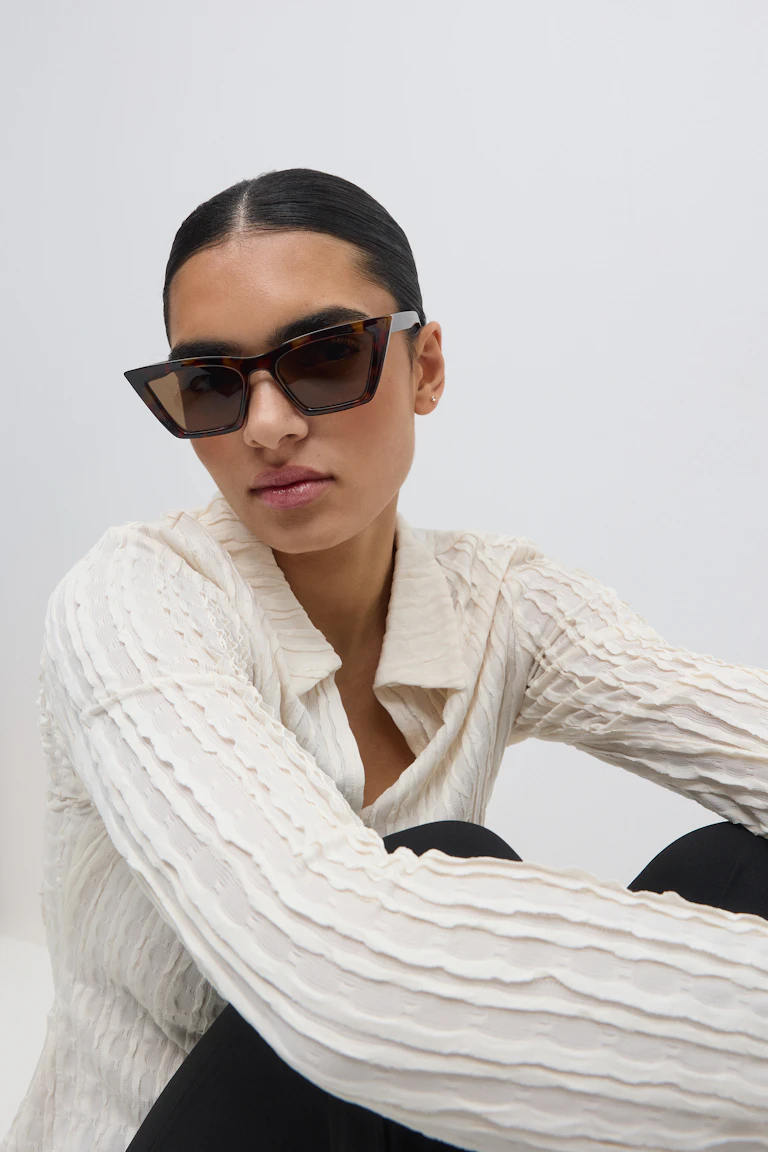 Een cat-eye zonnebril met schildpadmotief; een crèmekleurige blouse met lange mouwen en verticale rimpeldetails; een zwarte getailleerde broek.
