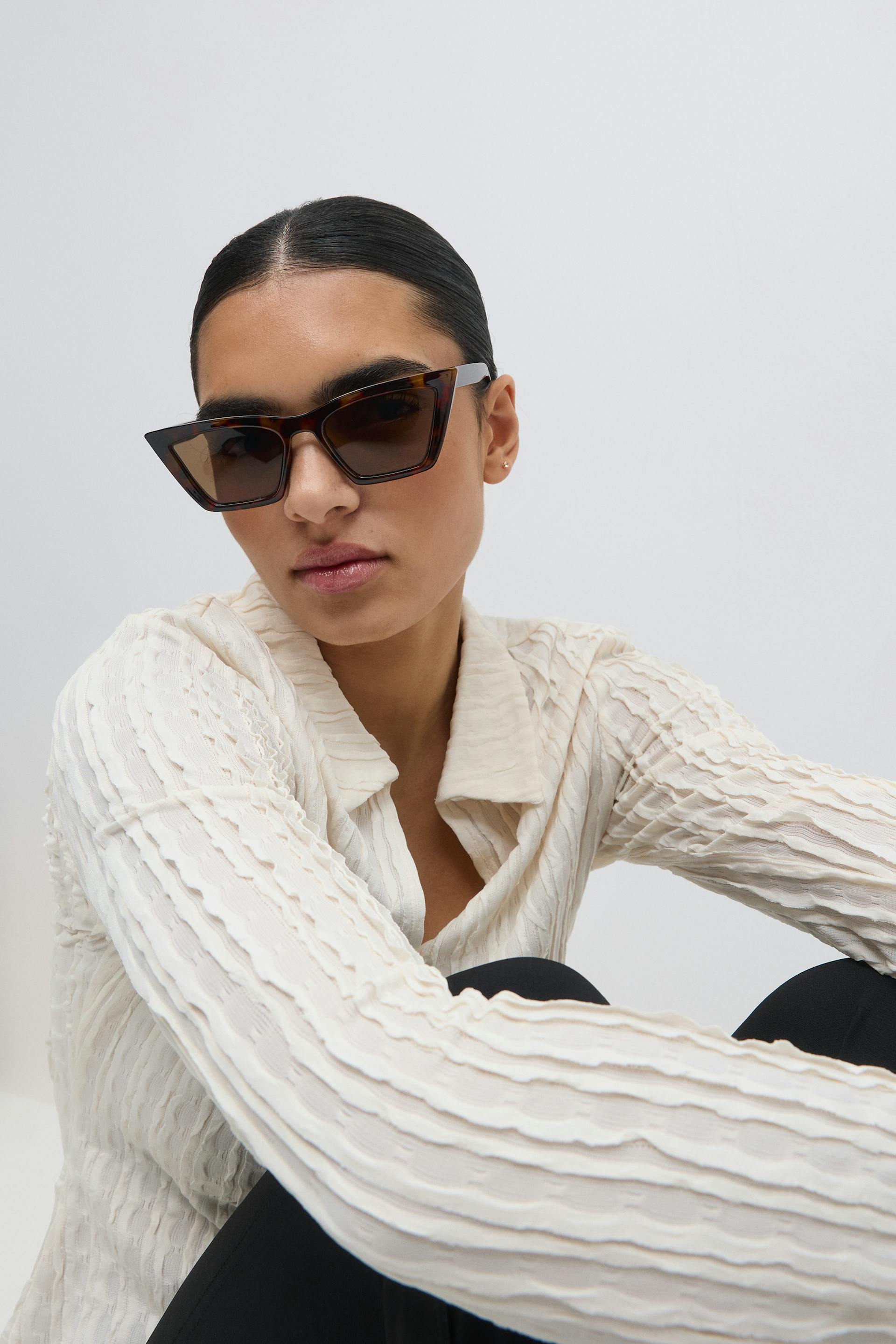 Tortoiseshell cat-eye sunglasses; cream textured long-sleeve collared blouse with vertical ripple detailing; black tailored trousers.