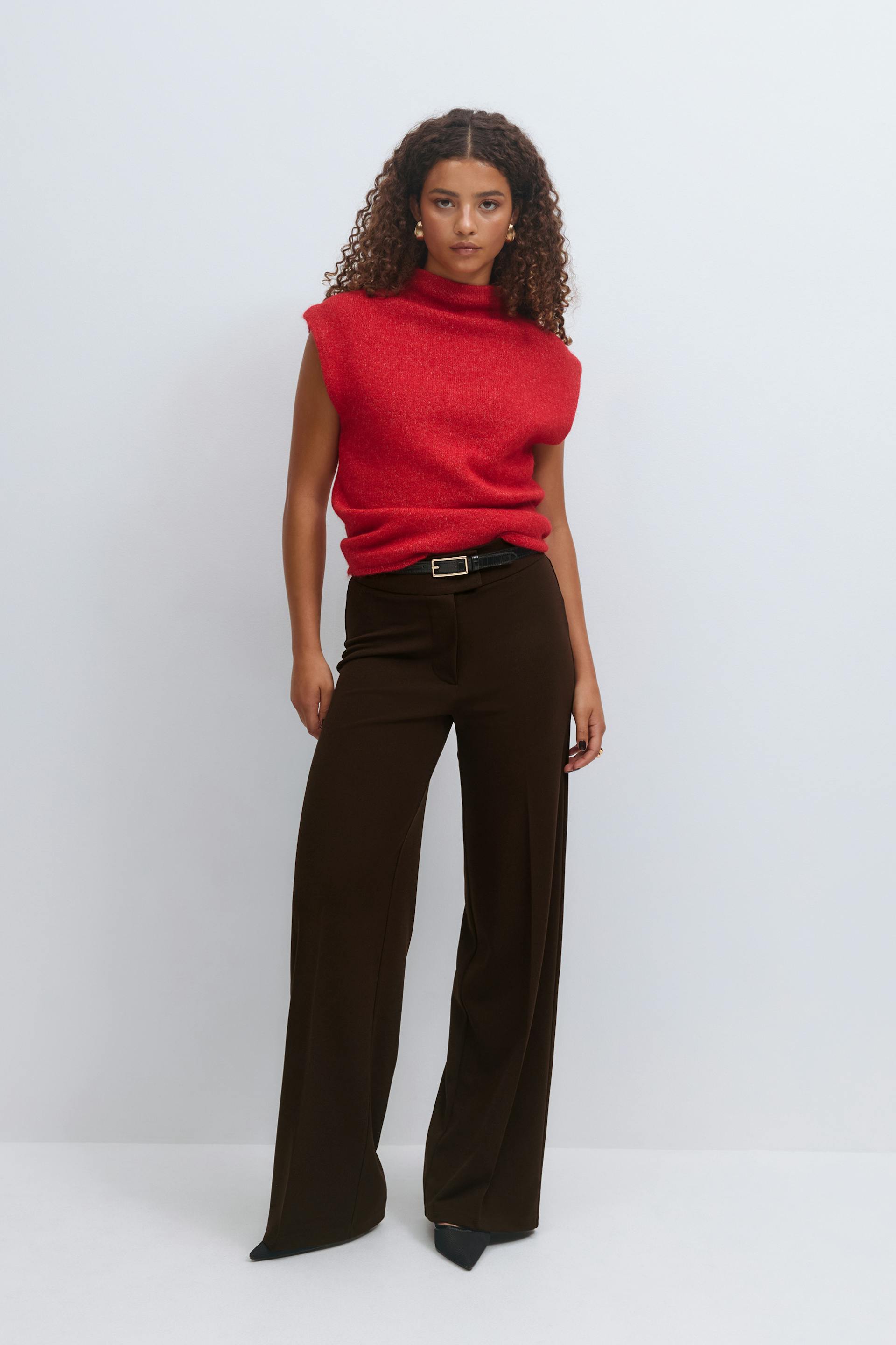 Sleeveless red mock‑neck knit top paired with high‑waisted dark brown wide‑leg trousers, a black belt with gold buckle, and black pointed‑toe heels.