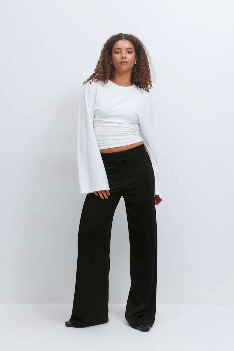 Witte top met lange mouwen en ruches in de taille, uitlopende mouwen, gecombineerd met een zwarte broek met hoge taille, wijde pijpen en zwarte schoenen met spitse tenen.