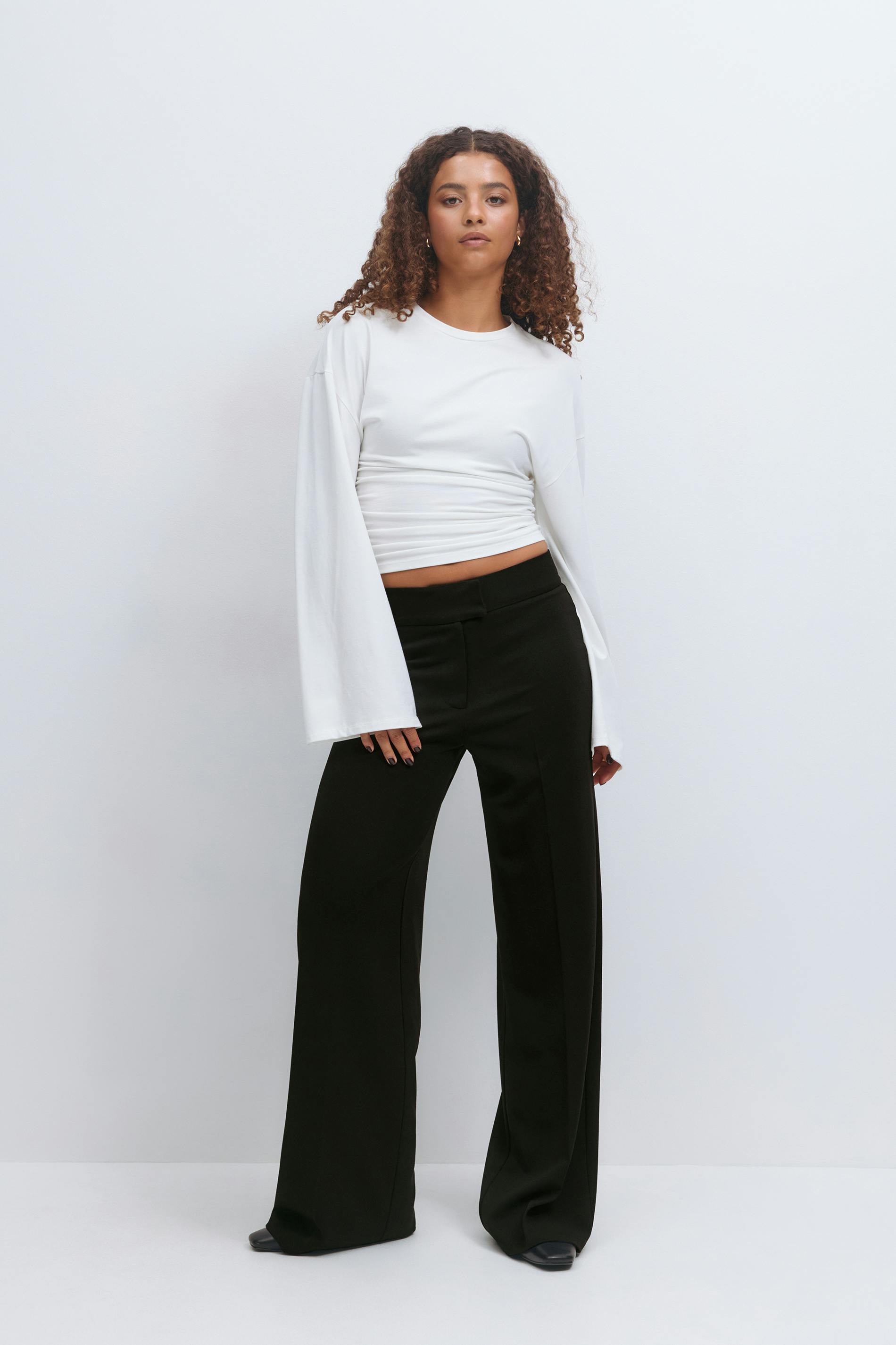 White long-sleeve cropped top with ruched waist and flared sleeves, paired with high-waisted black wide-leg trousers and black pointed-toe shoes.