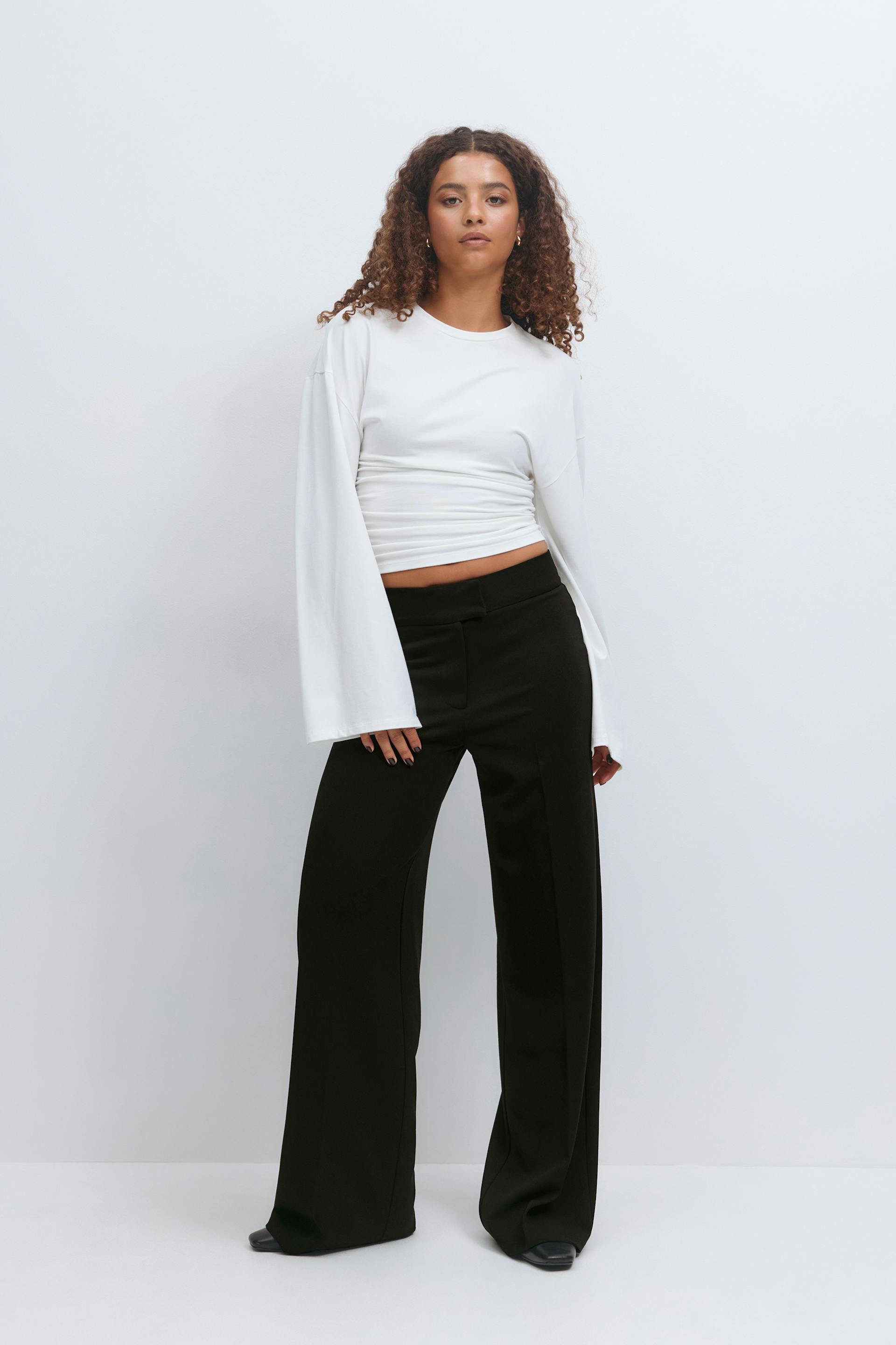 White long-sleeve cropped top with ruched waist and flared sleeves, paired with high-waisted black wide-leg trousers and black pointed-toe shoes.