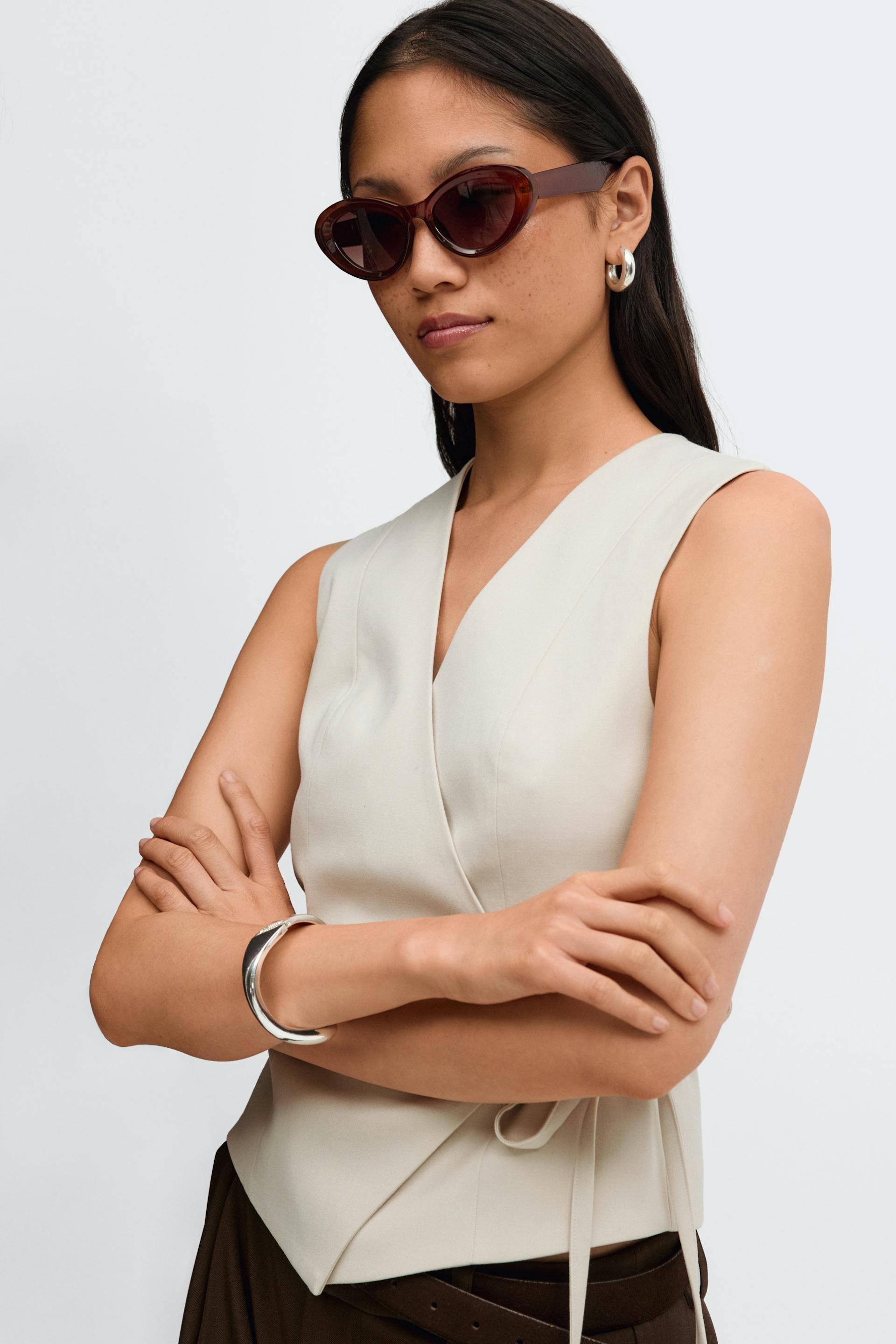 Ermeløs, beige topp med V-utringning og slips i siden, mørkebrune bukser med belte, kattesolbriller i skilpaddeskjell, øreringer i sølv og armbånd i sølv.