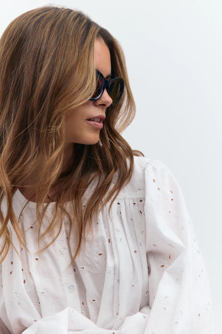 Hvid broderie anglaise-bluse med rynket bærestykke og puffede lange ærmer, sat sammen med sorte oversized cat-eye-solbriller.