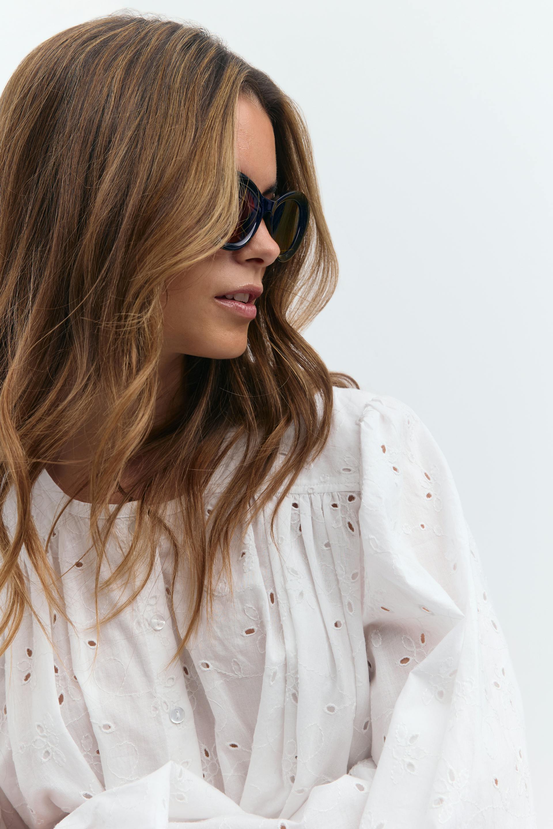 Valkoinen broderie anglaise -pusero, jossa on kerätty päärme ja pitkät paisutetut hihat, yhdistettynä mustiin ylimitoitettuihin kissansilmä-aurinkolaseihin.