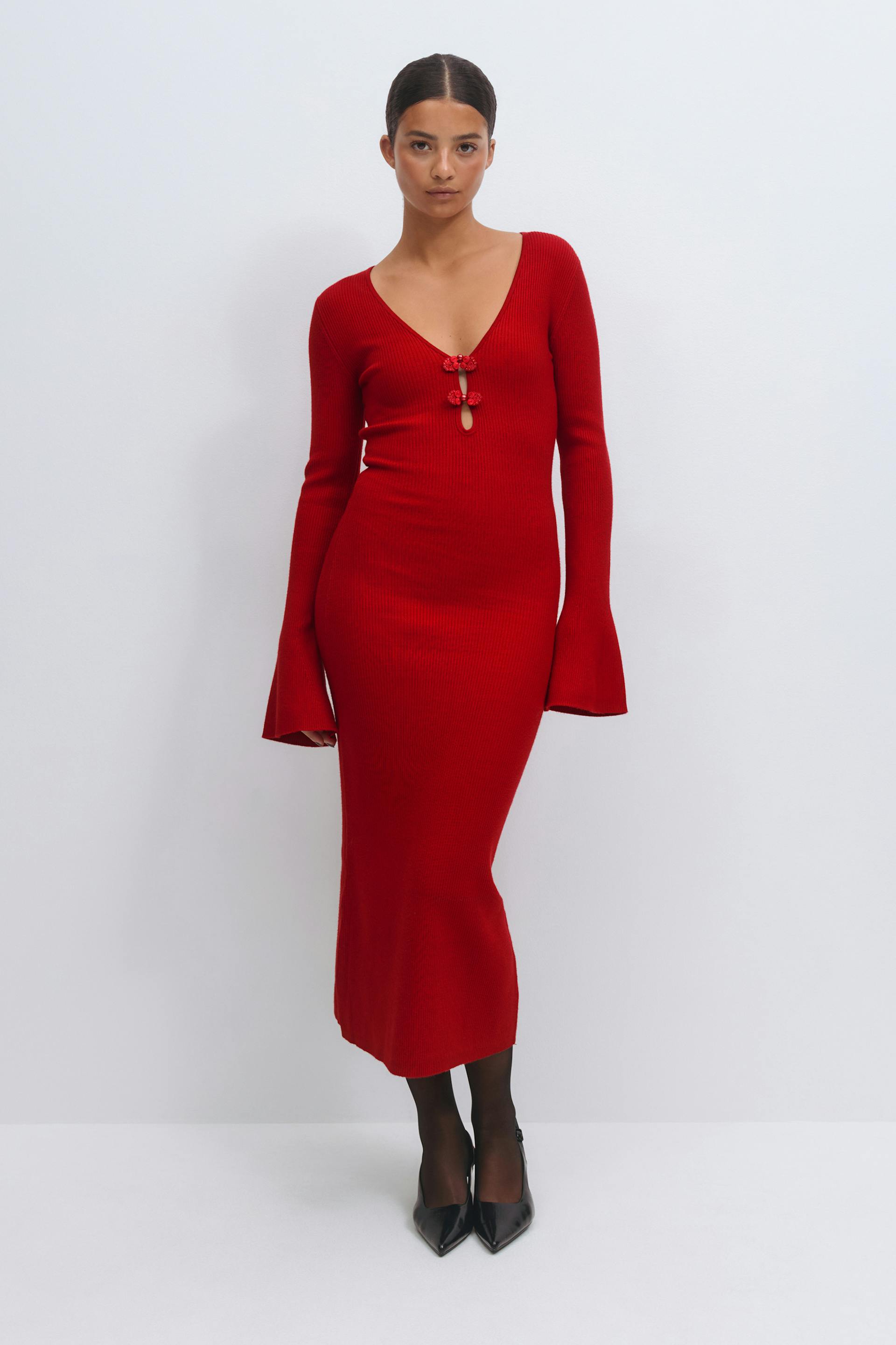 Rotes, langärmeliges Midikleid aus Rippstrick mit tiefem V-Ausschnitt, zwei dekorativen Knotenverschlüssen an der Brust und ausgestellten Manschetten, kombiniert mit einer schwarzen durchsichtigen Strumpfhose und schwarzen spitzen Absätzen.