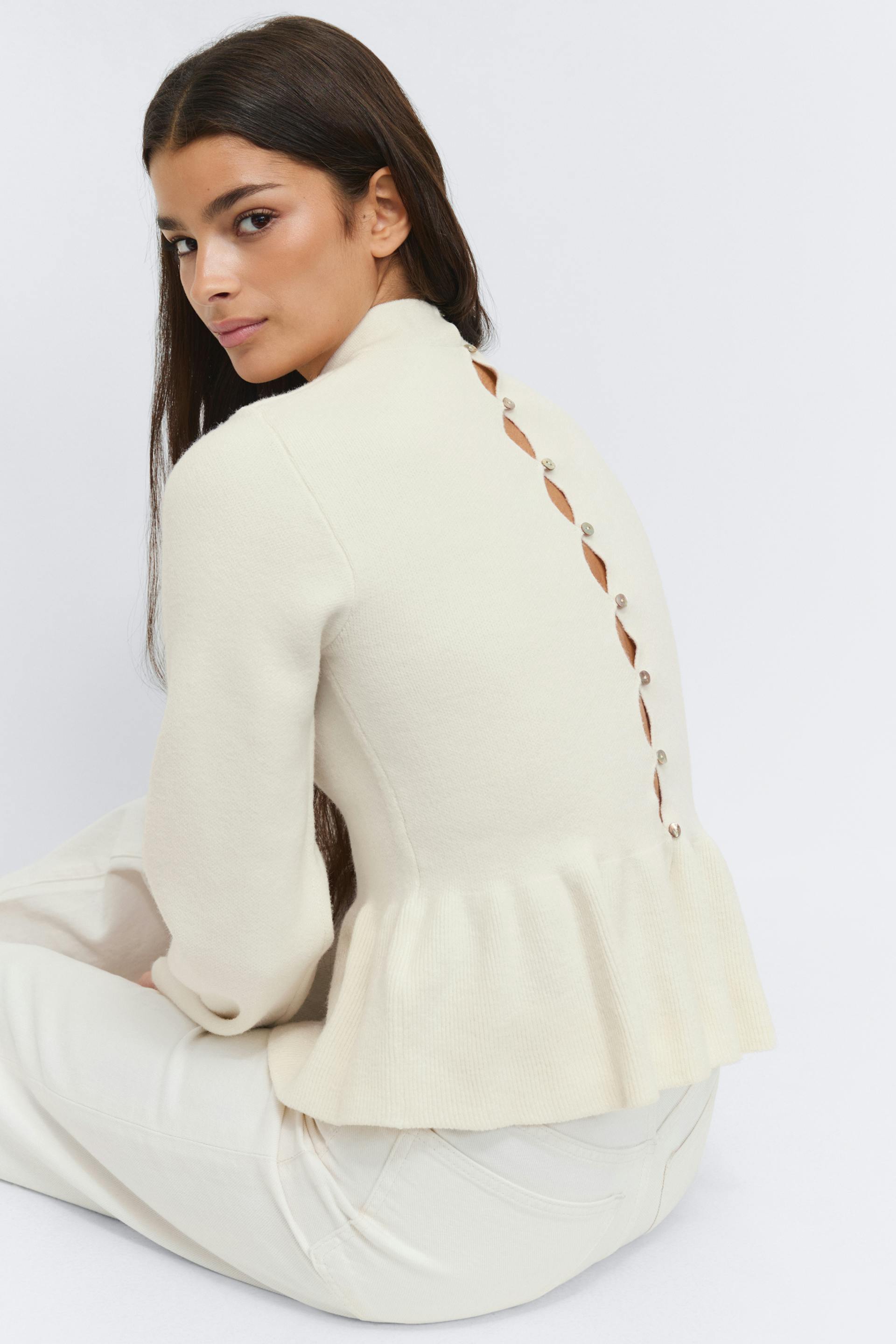 Ivoorkleurige gebreide mock-hals trui met lange mouwen, peplum zoom en een verticale rij knoopjes met druppelvormige uitsnijdingen aan de achterkant, gecombineerd met een crèmekleurige broek met hoge taille.