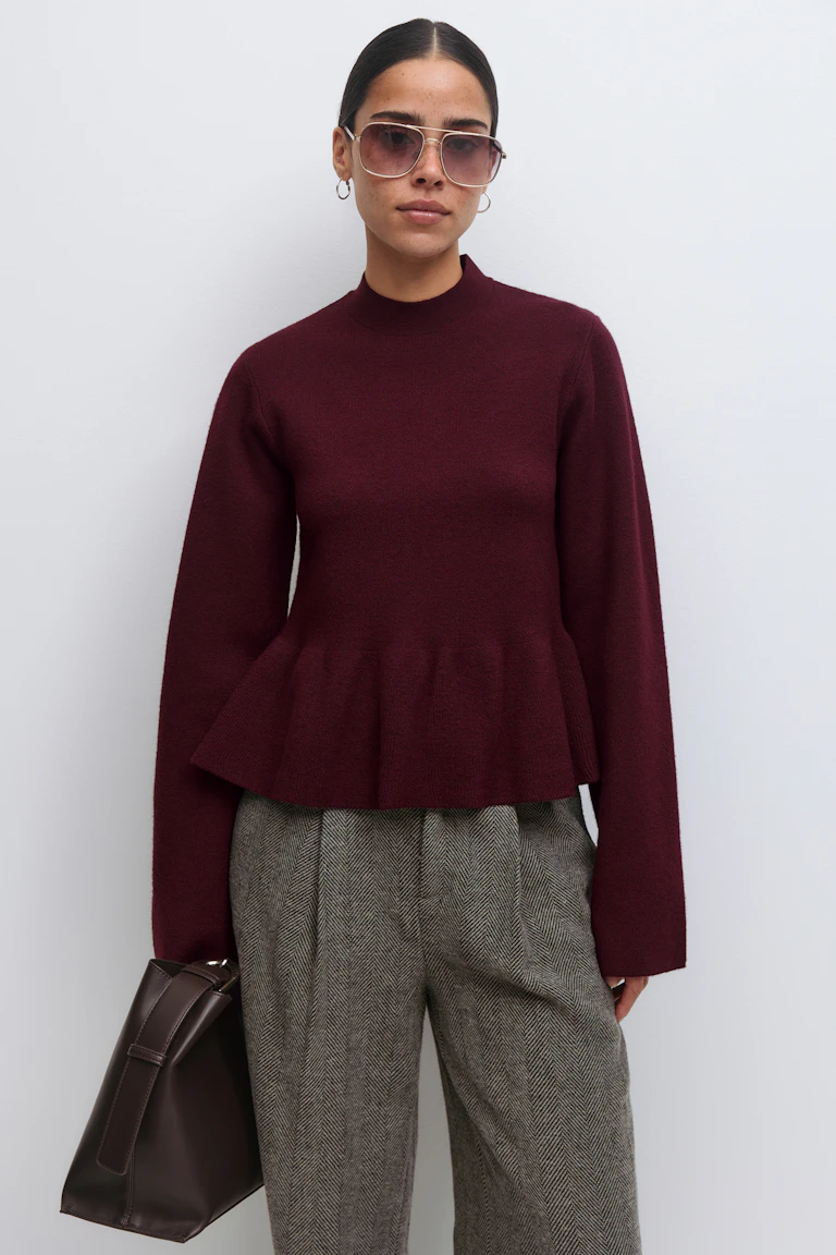 Burgundy mock‑neck peplum sweater with long sleeves; grey herringbone pleated trousers; dark brown leather tote; gold hoop earrings and aviator sunglasses.