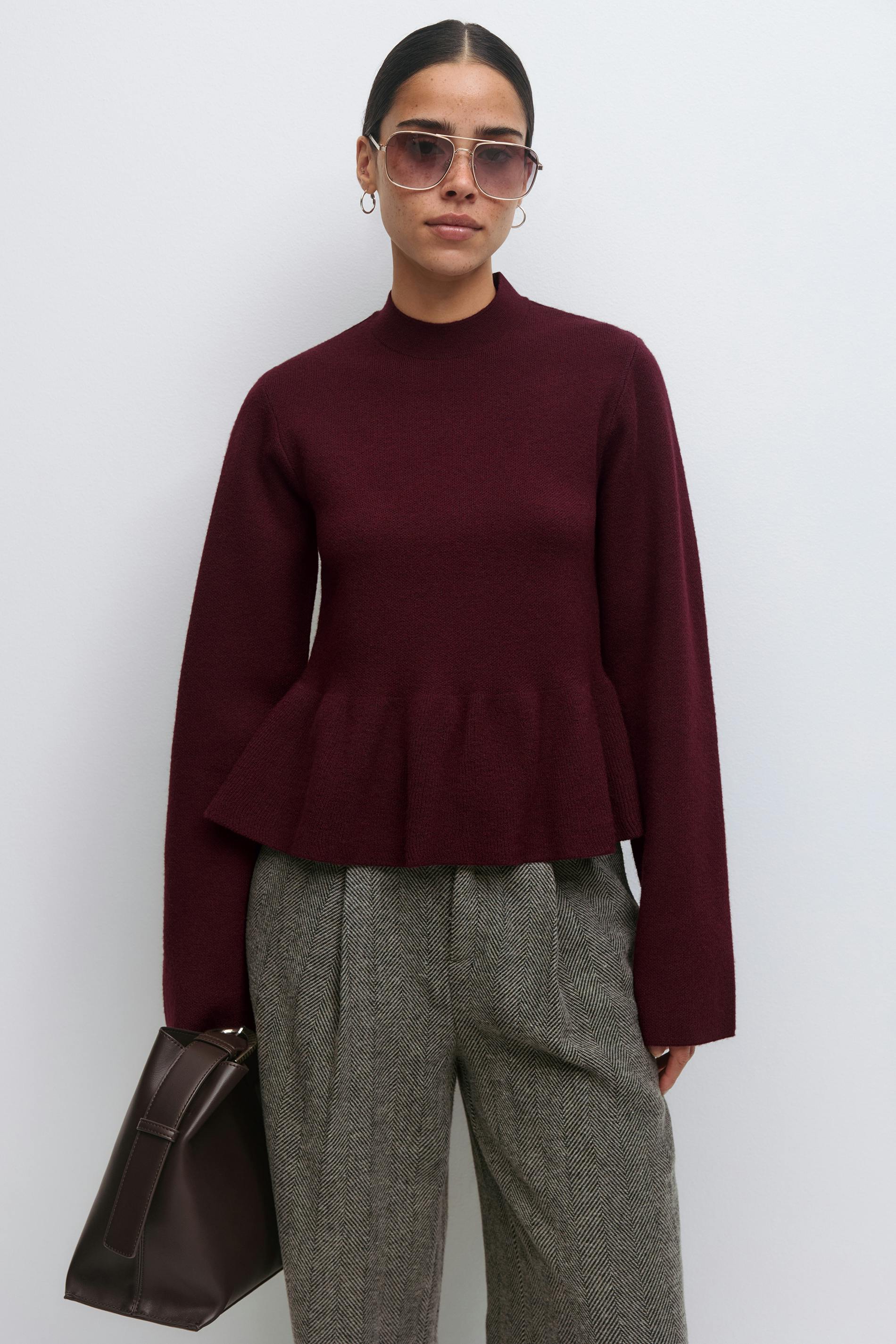 Burgunderfarbener Pullover mit Schößchen und langen Ärmeln; graue Hose mit Fischgrätenfalten; dunkelbraune Ledertasche; goldene Ohrringe und Pilotensonnenbrille.