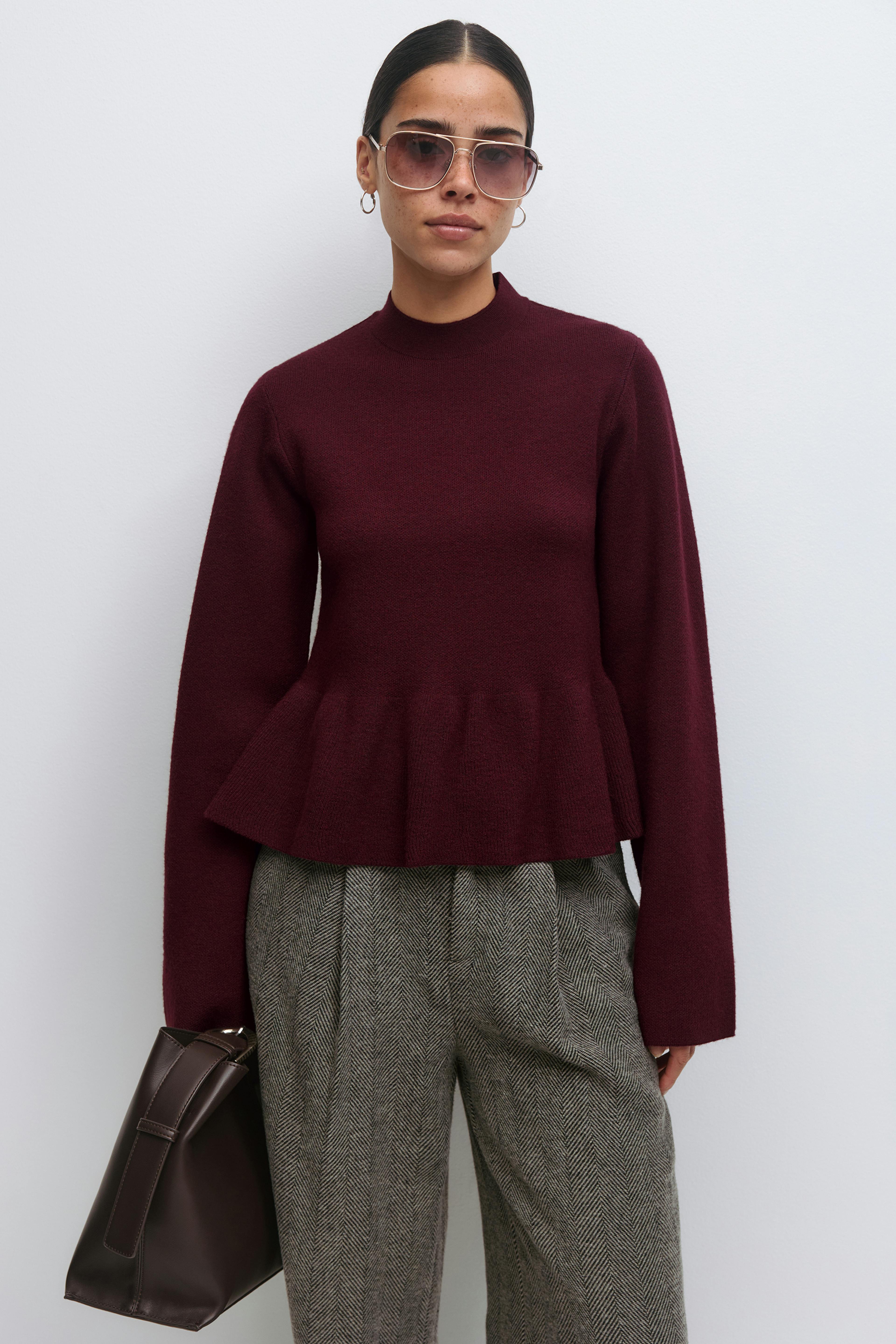 Bourgognefarvet peplum-sweater med mock-neck og lange ærmer; grå sildebensplisserede bukser; mørkebrun lædertaske; guldøreringe og pilotsolbriller.