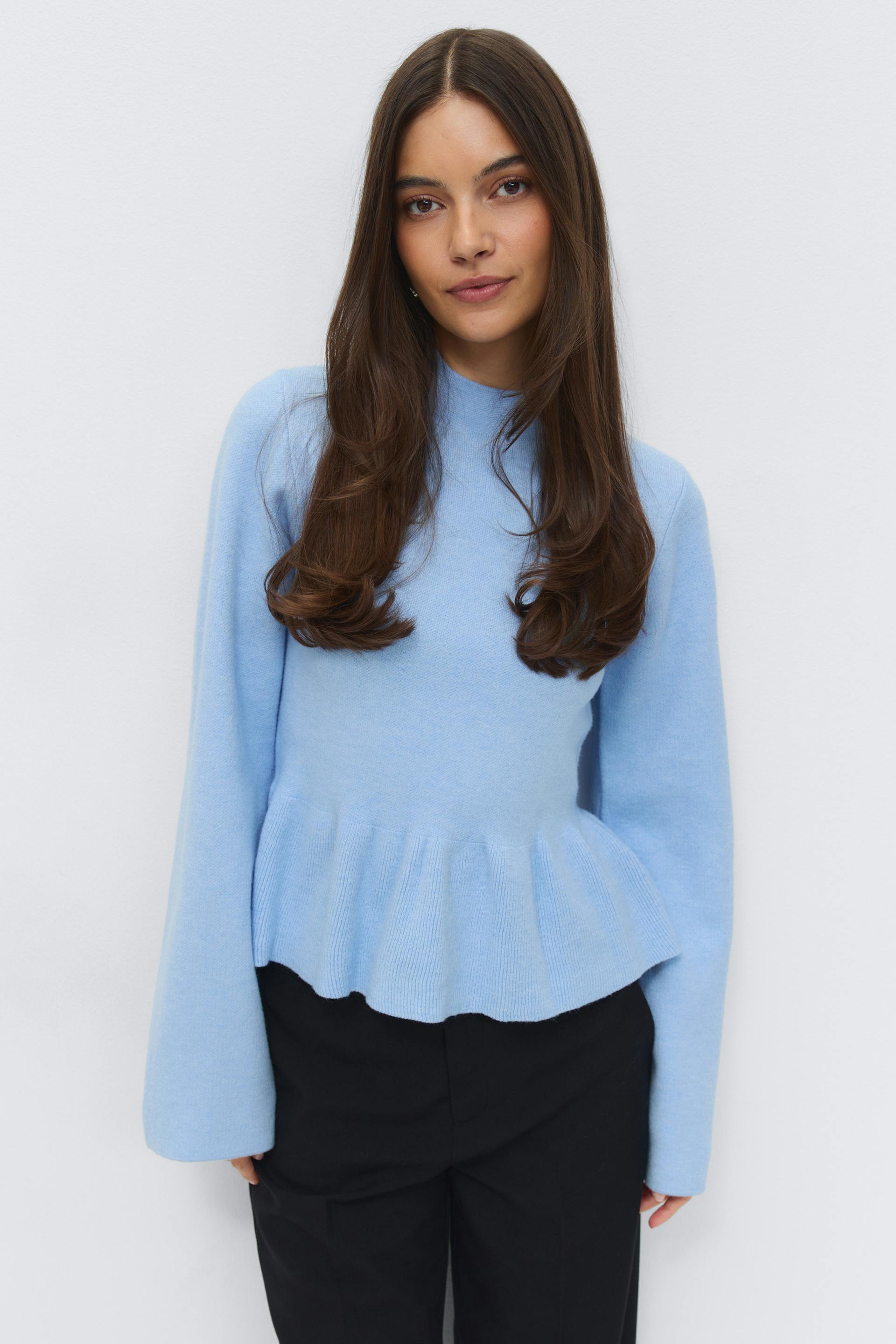 Light blue long-sleeve knit sweater with a mock neck and peplum hem, paired with black tailored trousers.