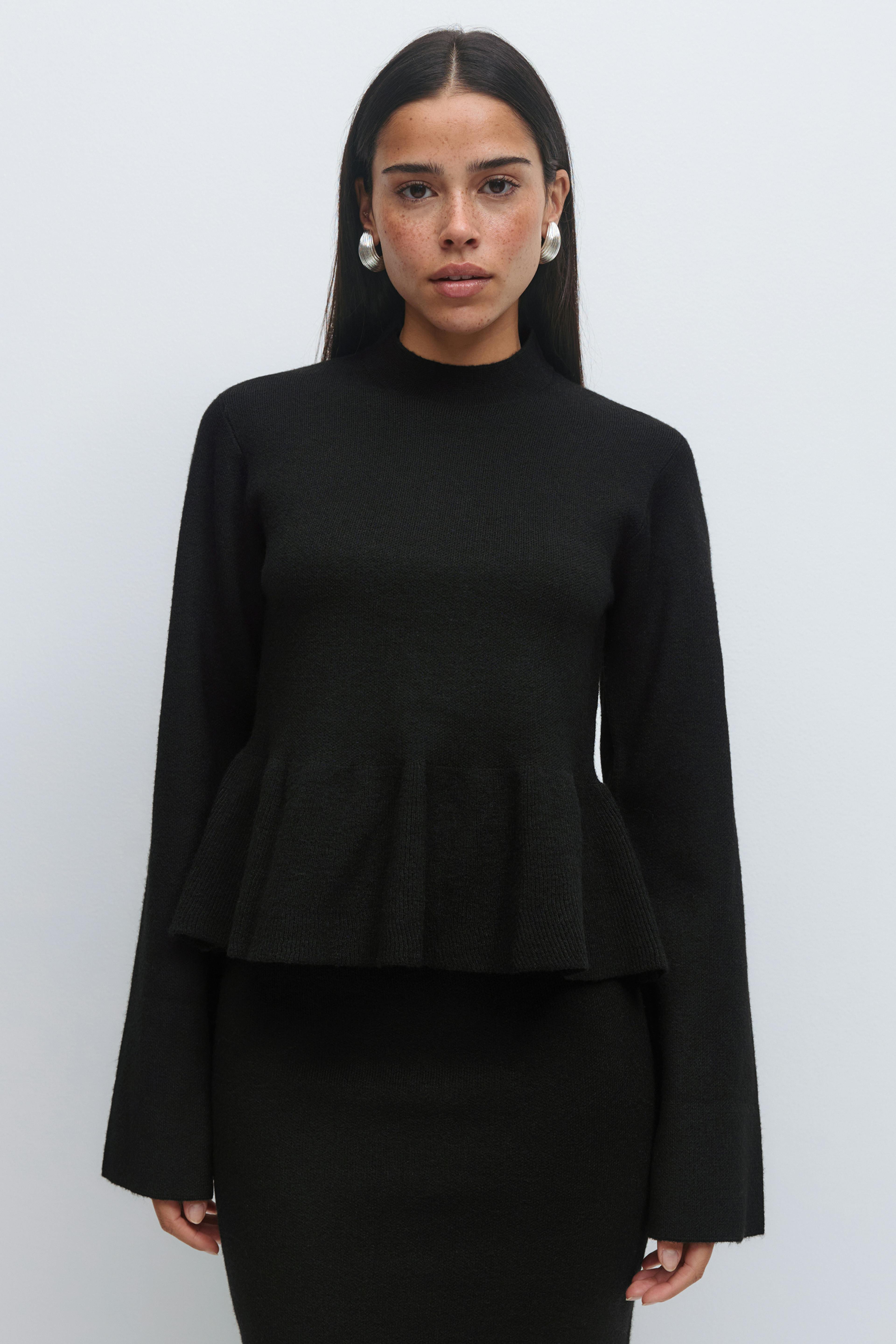 Musta pitkähihainen mock-neuletoppi, jossa on peplum-helma, yhdistettynä sovitettuun mustaan hameeseen ja suuriin hopeisiin renkaisiin korvakoruihin.