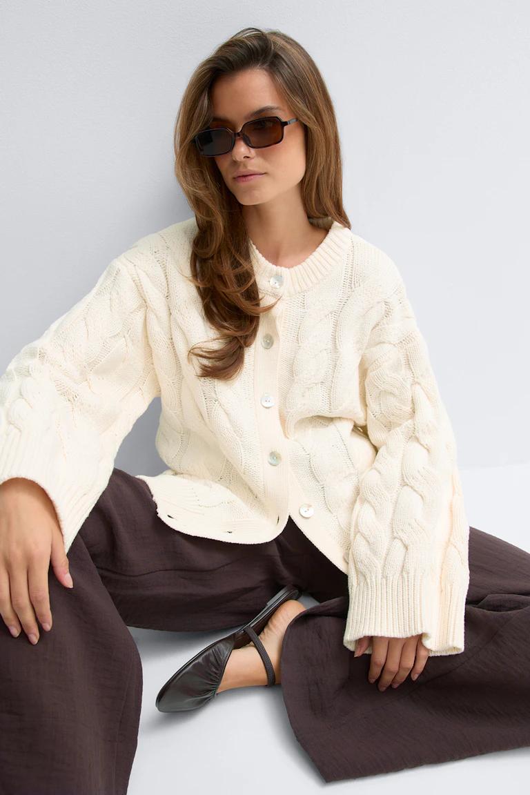 Elfenbensfarget kabelstrikket cardigan med knappefront, oversized passform og ribbestrikkede mansjetter, kombinert med mørkebrune bukser med vide ben og spisse slingback-flater i mørkt skinn, stylet med rektangulære, brune solbriller.
