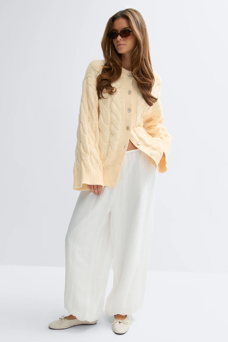 Übergroße cremefarbene Strickjacke mit Knopfleiste, eine weite weiße Hose, beigefarbene Ballettschuhe mit Schleife und eine dunkle rechteckige Sonnenbrille.