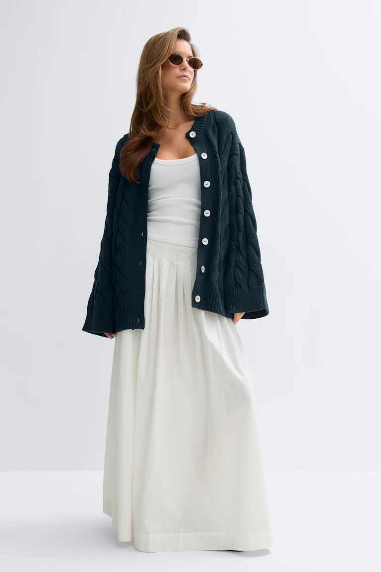 Mörk kabelstickad oversized genomknäppt cardigan med stora vita knappar, vitt figursytt linne, vit plisserad golvlång kjol och små ovala solglasögon.