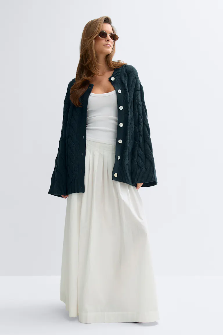 Mørk kabelstrikket oversized gennemknappet cardigan med store hvide knapper, hvid tætsiddende tanktop, hvid plisseret gulvlang nederdel og små ovale solbriller.