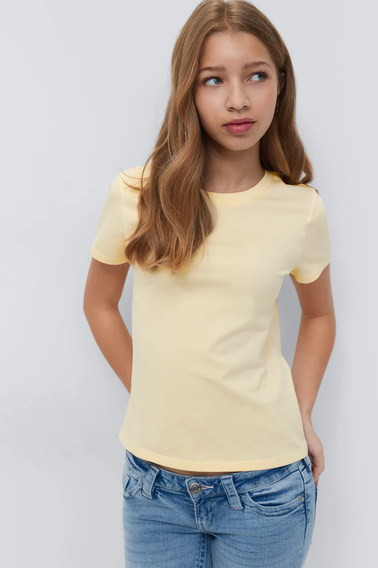Hellgelbes, kurzärmeliges T-Shirt mit Rundhalsausschnitt, kombiniert mit hellblauen, halbhohen Jeans mit Gürtelschlaufen und Knopfverschluss.
