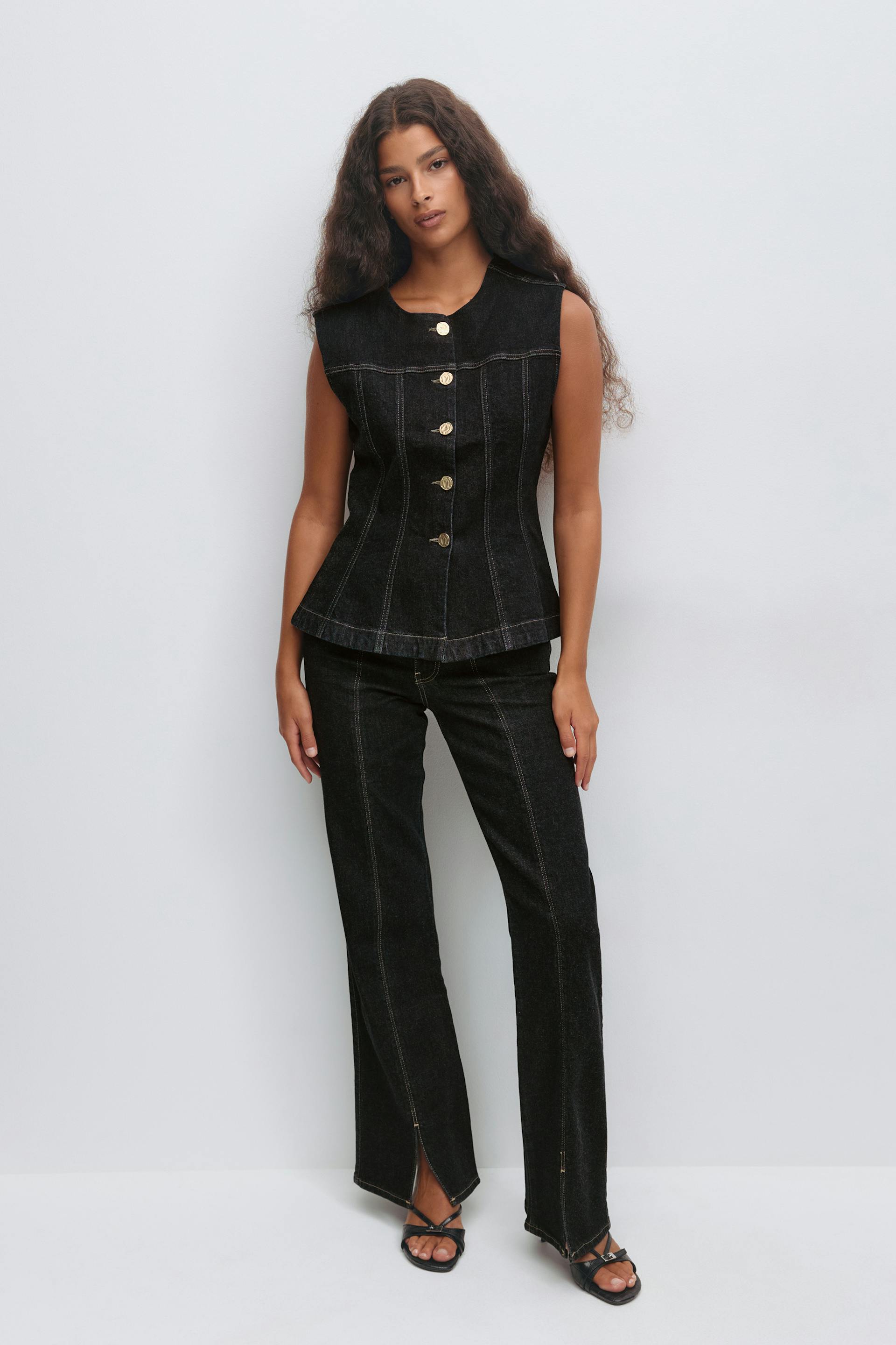 Sleeveless black denim fitted button-front top with gold buttons and contrast seam detailing, paired with matching black straight-leg denim trousers with central front seams and front hem slits, and black strappy heeled sandals.