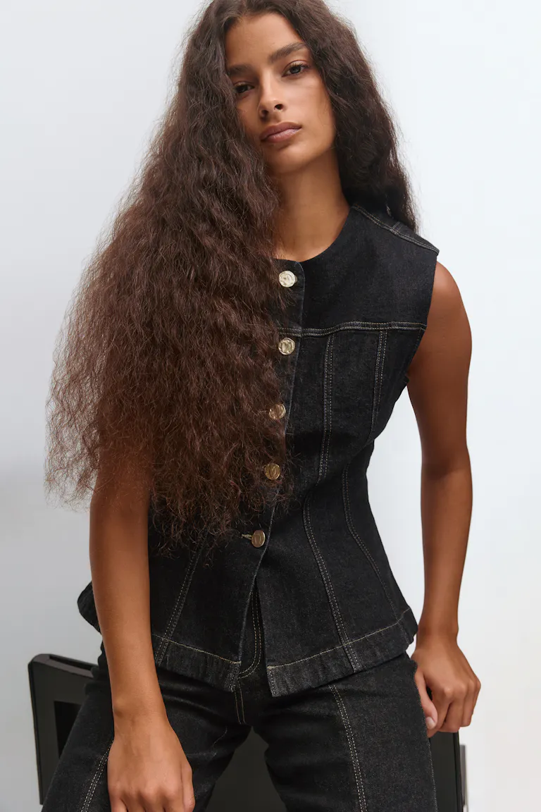 Ärmellose schwarze Jeansweste mit goldfarbenen Knöpfen und kontrastierenden Steppnähten, kombiniert mit einer passenden schwarzen Jeans.
