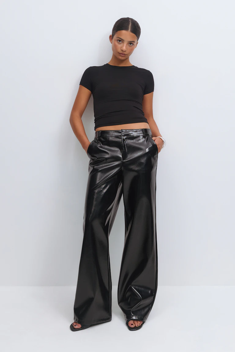 Er trägt ein schwarzes, kurzärmeliges, tailliertes Oberteil, eine schwarze, hochtaillierte, glänzende Kunstlederhose mit weitem Bein, schwarze, offene Sandalen und einen silbernen Armreif.