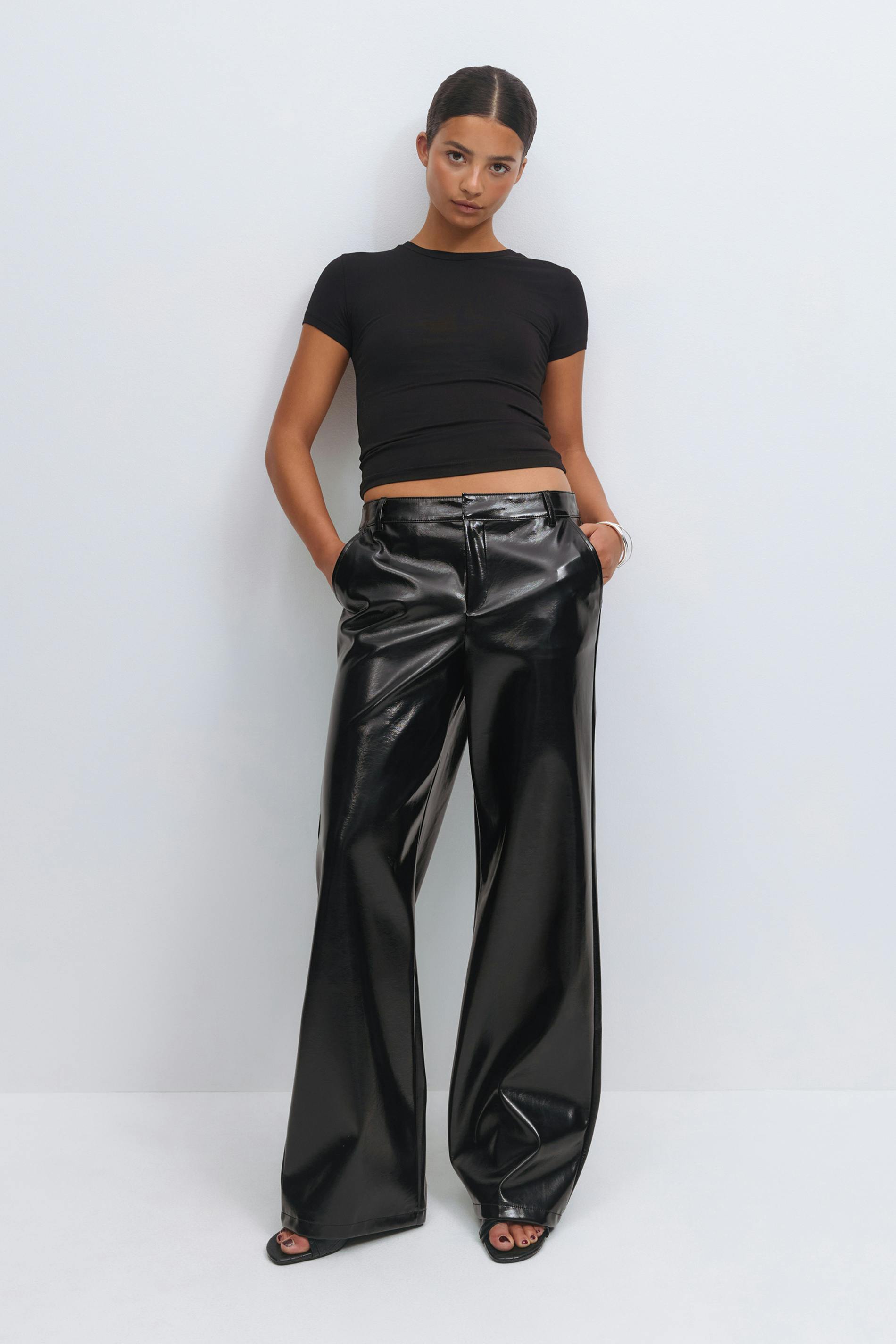 Wearing a fitted black short-sleeve cropped top, high-waisted glossy black wide-leg faux-leather trousers, black open-toe sandals and a silver bangle.