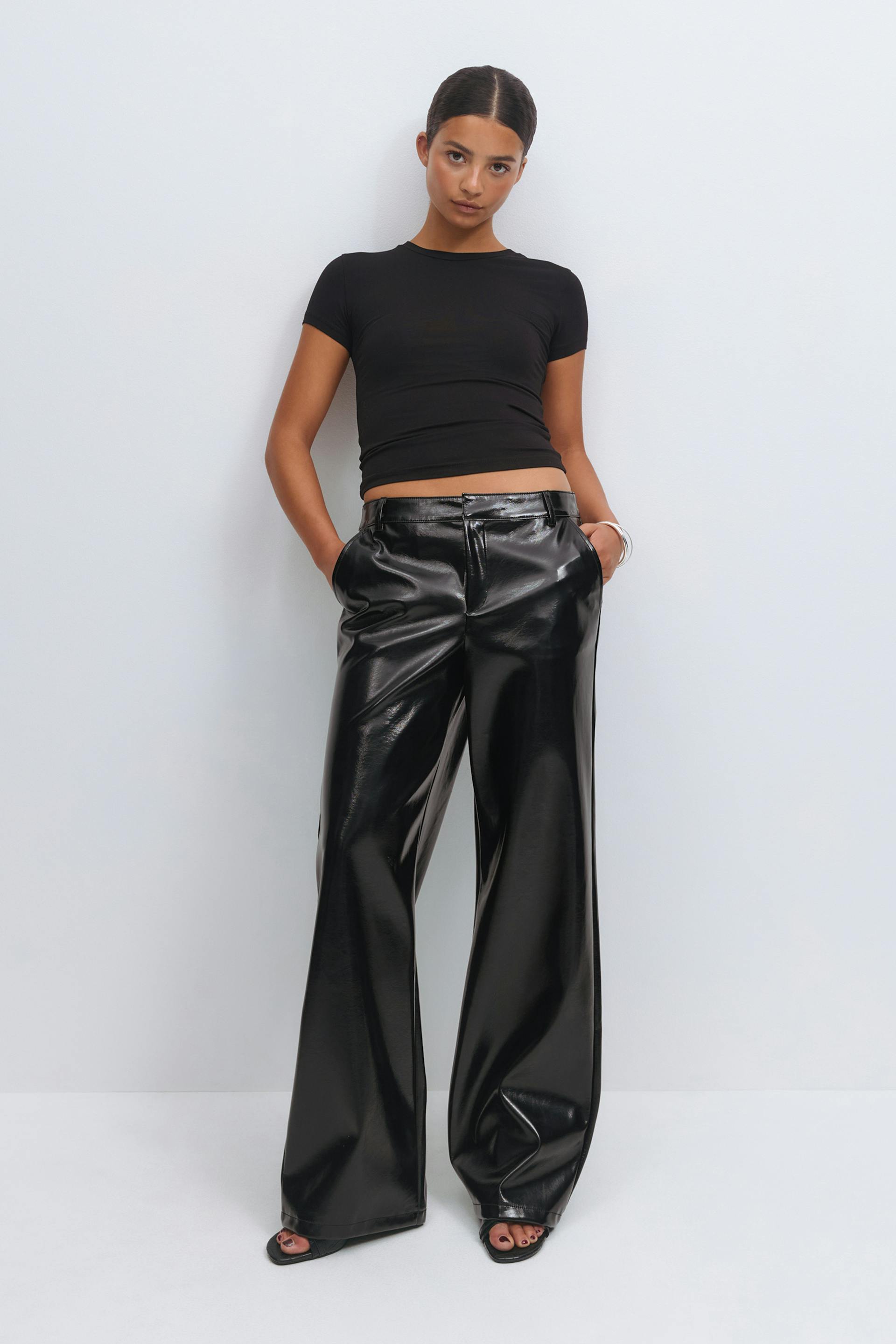 Wearing a fitted black short-sleeve cropped top, high-waisted glossy black wide-leg faux-leather trousers, black open-toe sandals and a silver bangle.