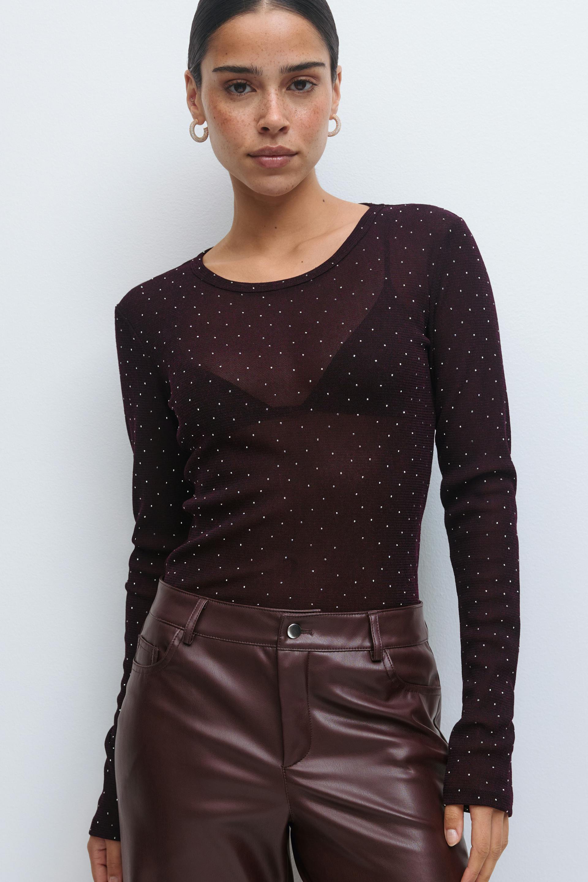 Durchsichtiges pflaumenfarbenes, langärmeliges Strickoberteil mit kleinen Metallic-Punkten über einem schwarzen Bralette; burgunderrote Lederhose mit hoher Taille; goldene Ohrringe.